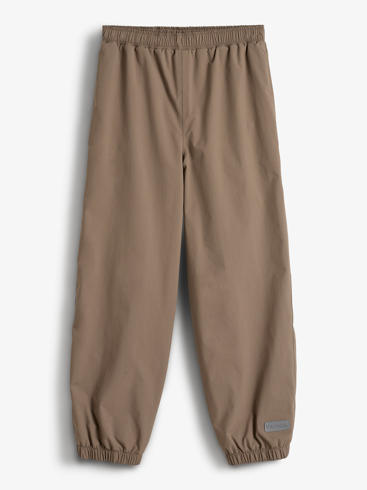 Bukser med elastisk talje og ben, der hviler fladt mod en lys baggrund. De er ensfarvede brune med en lille mærkat ved anklen med teksten "Polarn O. Pyret".