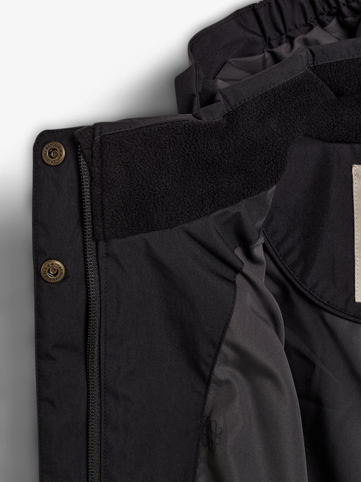 Jakke med sort materiale og krave foret med fleece, placeret indendørs. Der er en messingknap ved siden af lynlåsen, der åbner jakken.