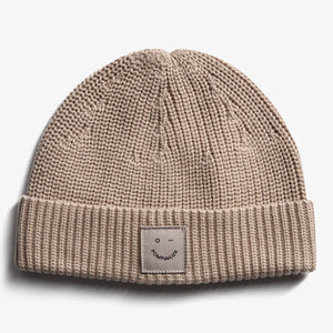En beige strikket hue med et ombukket kant. På forsiden er der en mærkat med teksten "pOMPdeLUX" og en simpel ansigtsskitse. Baggrunden er helt hvid.