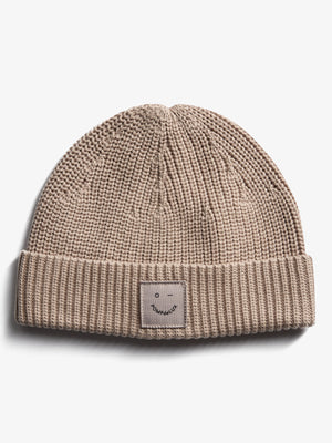 En beige strikket hue med et ombukket kant. På forsiden er der en mærkat med teksten "pOMPdeLUX" og en simpel ansigtsskitse. Baggrunden er helt hvid.