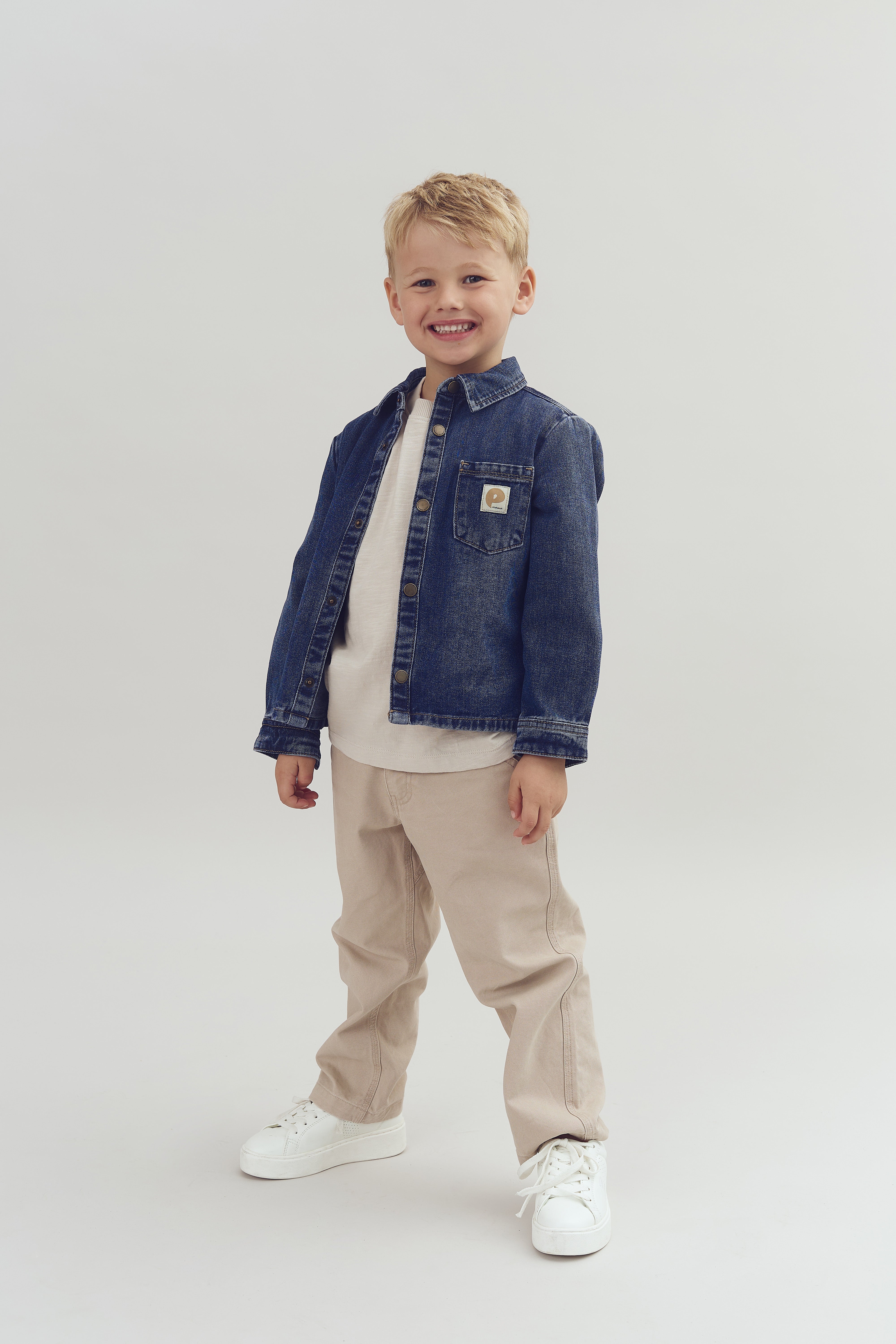 A smiling young child stands confidently, wearing a denim jacket, beige pants, and white sneakers, against a plain, light-colored background.