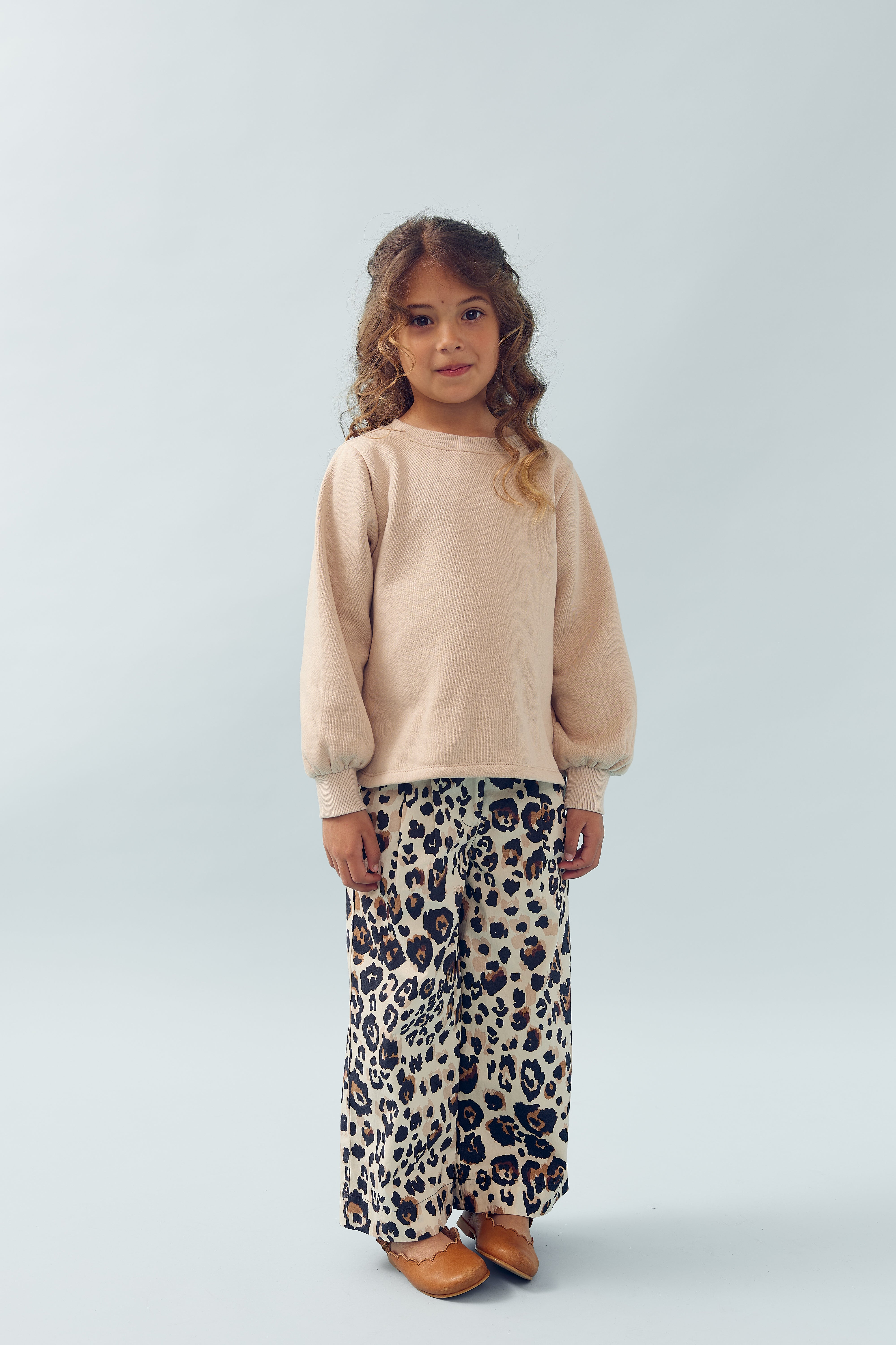 Et barn står iført en beige trøje og leopardmønstrede bukser i en lys, minimalistisk baggrund. Hun ser direkte mod kameraet.