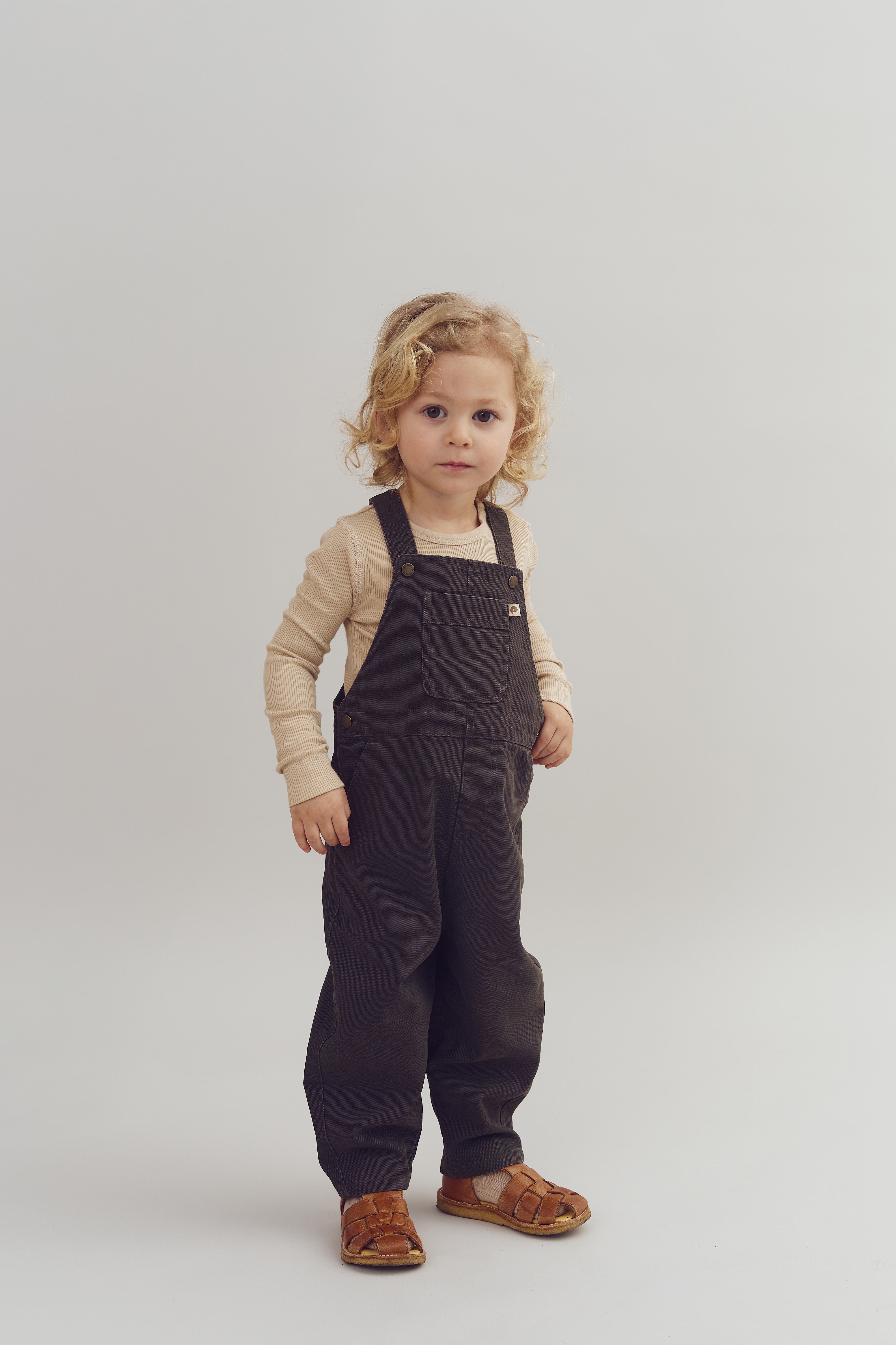 Et barn iført mørke overalls og lyse trøje står stille i et neutralt, lyst rum med en enkel baggrund. Barnet har blondt, krøllet hår og brune sandaler.