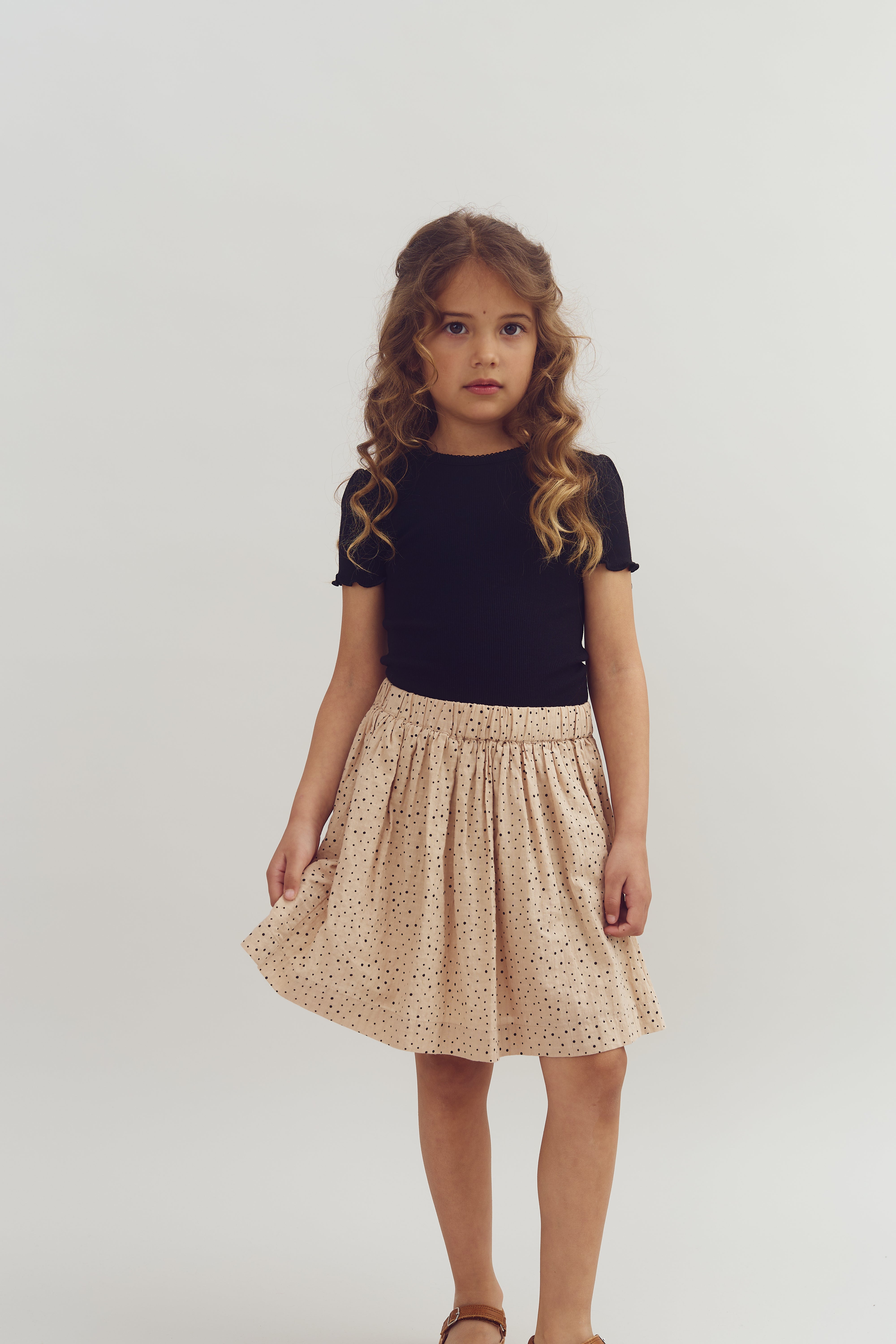 A child with long curly hair stands wearing a black shirt and a polka-dotted skirt, looking at the camera in a neutral expression against a plain white background.