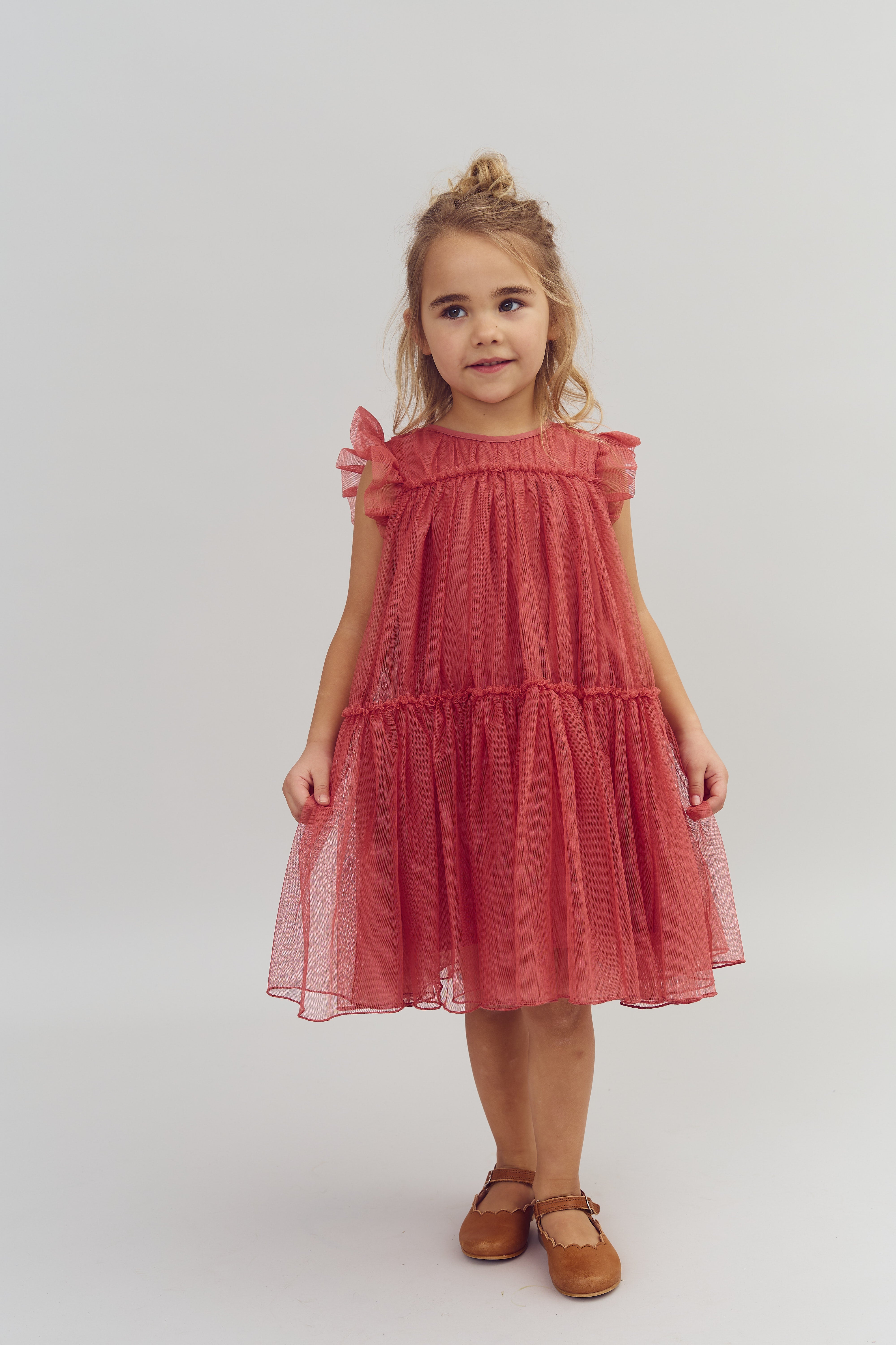 A young girl stands wearing a pink ruffled dress and brown shoes, smiling slightly. She is in a neutral, well-lit studio with a plain background.