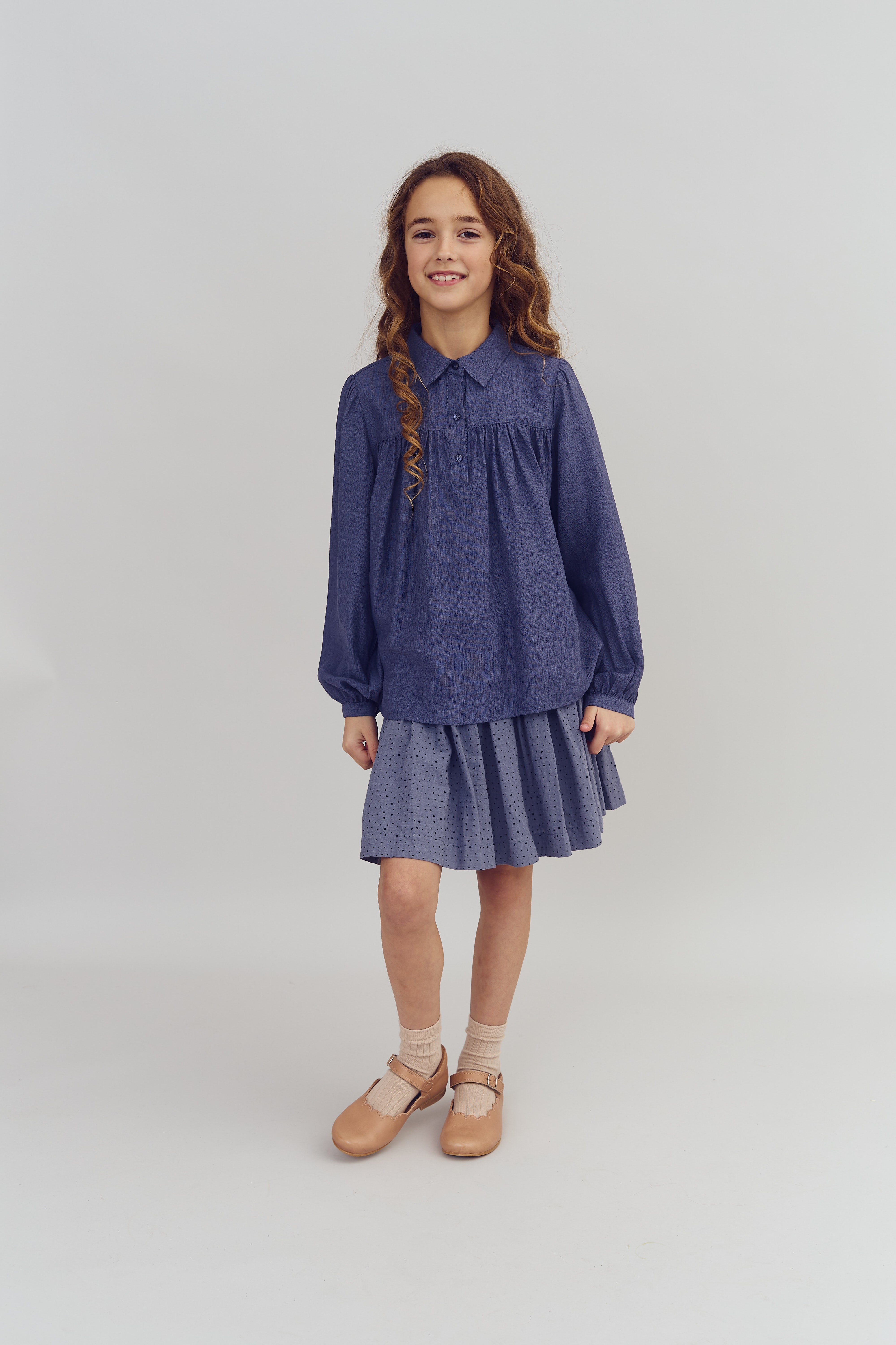 A young girl stands smiling, wearing a blue blouse, a pleated blue skirt, beige socks, and tan shoes. She is in a plain, neutral indoor setting with no text visible.