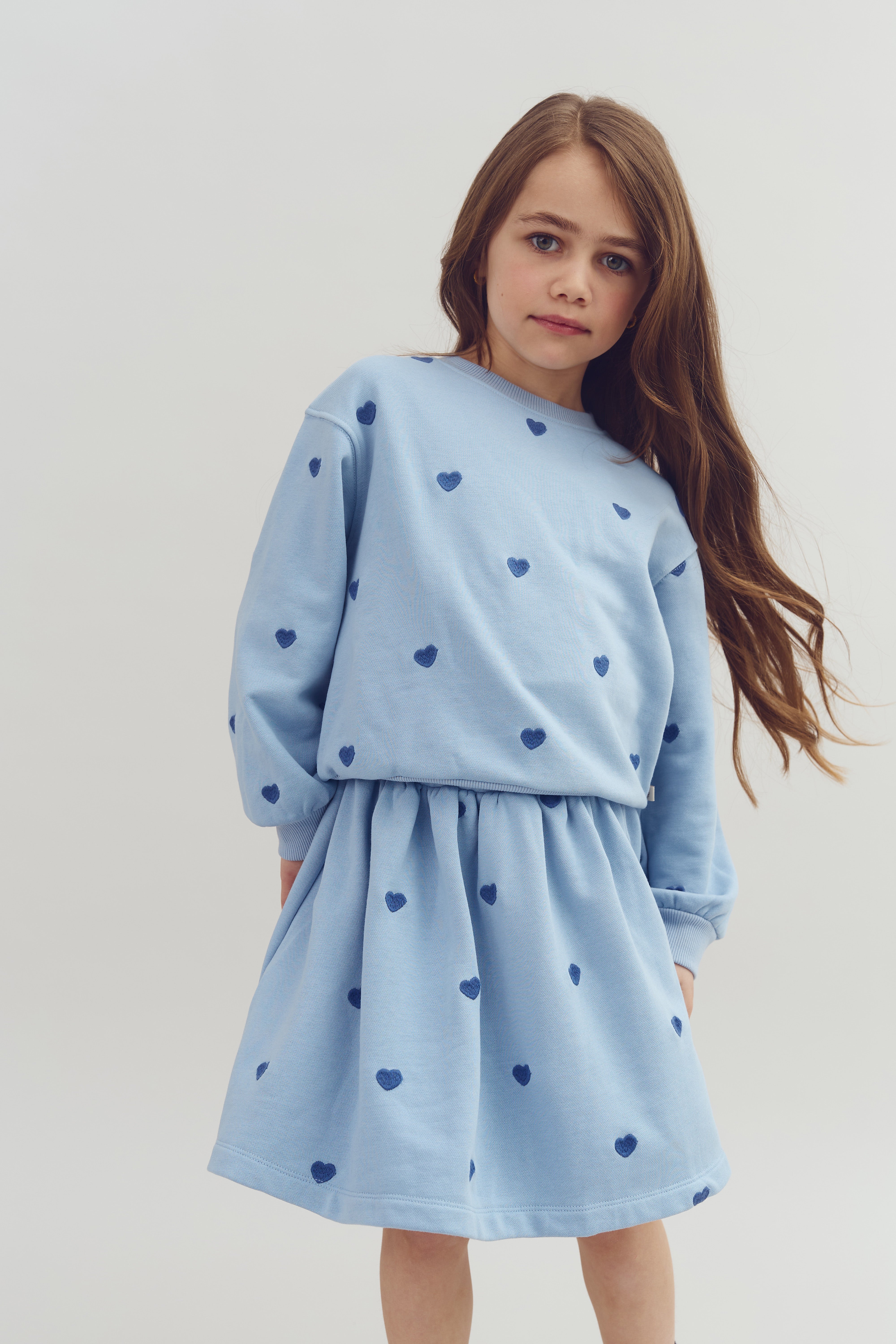 A girl in a blue dress with heart patterns stands confidently, hands in pockets, against a plain background. Her long hair falls over her shoulder.