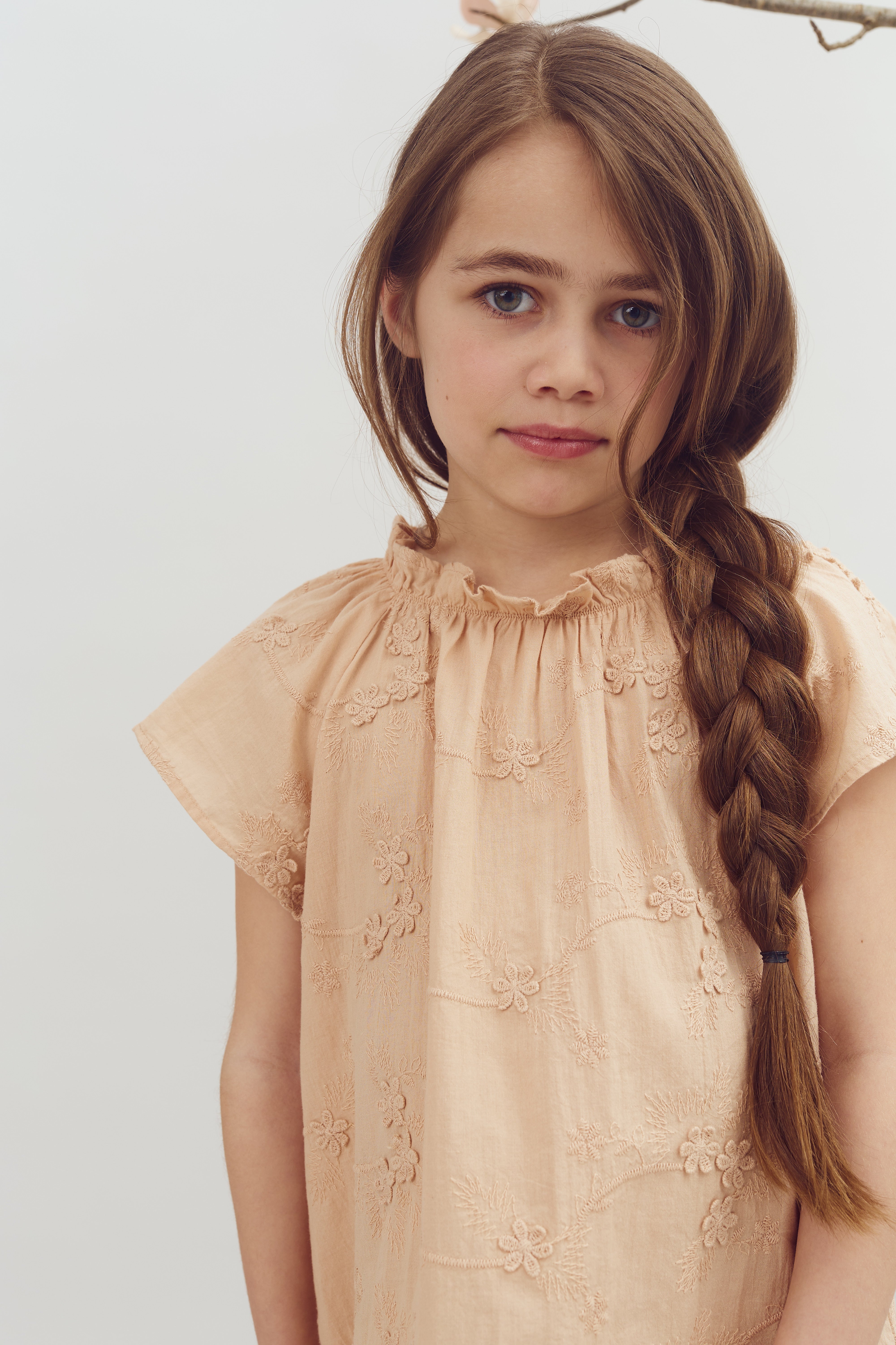 A young girl with braided hair stands, wearing a peach embroidered dress. She looks directly at the camera, set against a plain, light background.