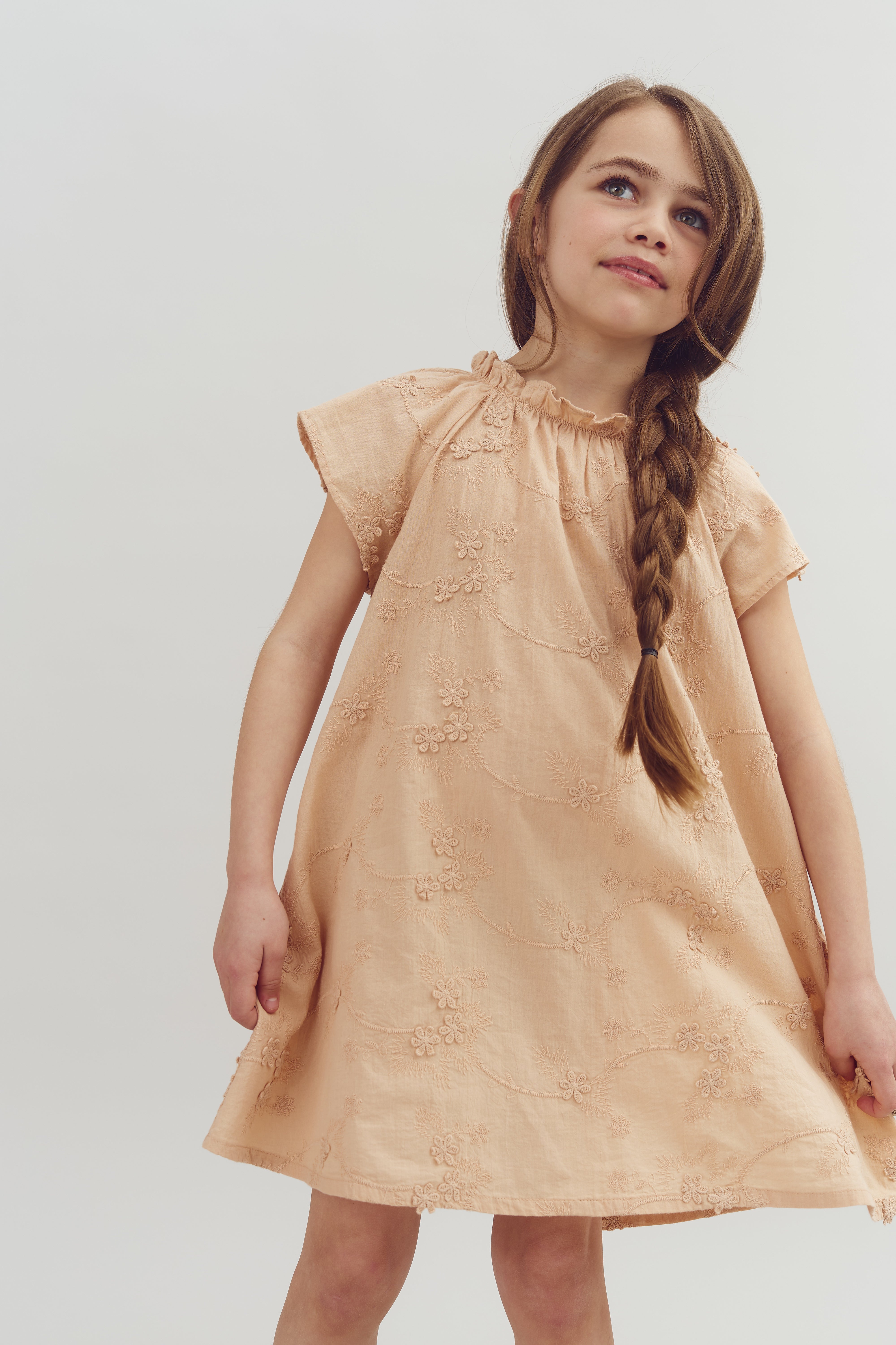 A child stands wearing a peach-colored dress with floral embroidery, her long hair braided, against a plain white background.
