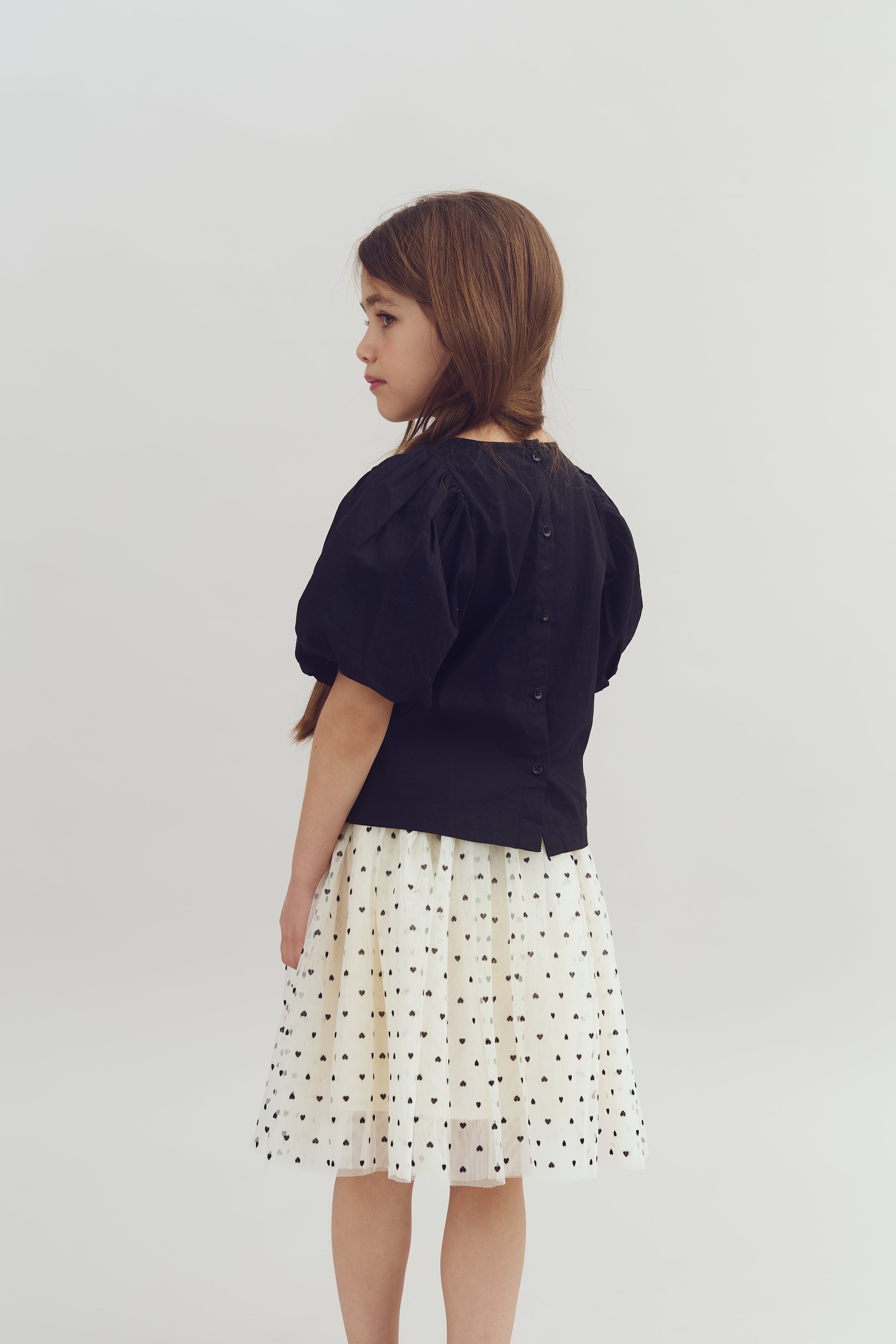 A young girl stands sideways, wearing a black top with buttons and a white skirt featuring small heart patterns, against a plain light background.