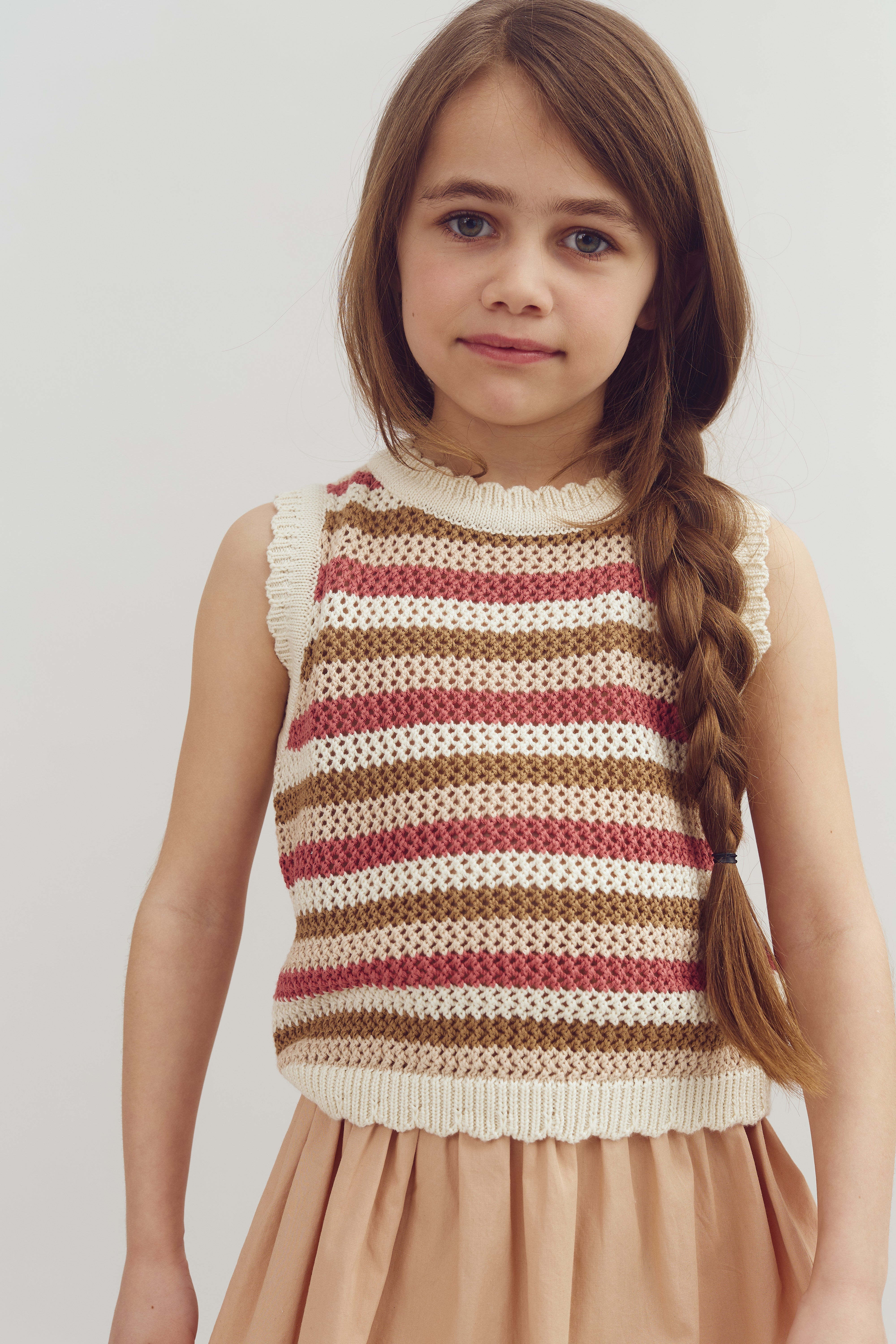 A young girl is standing with a calm expression, wearing a striped knit top and a beige skirt. Her long hair is styled in a braid, and the background is plain white.