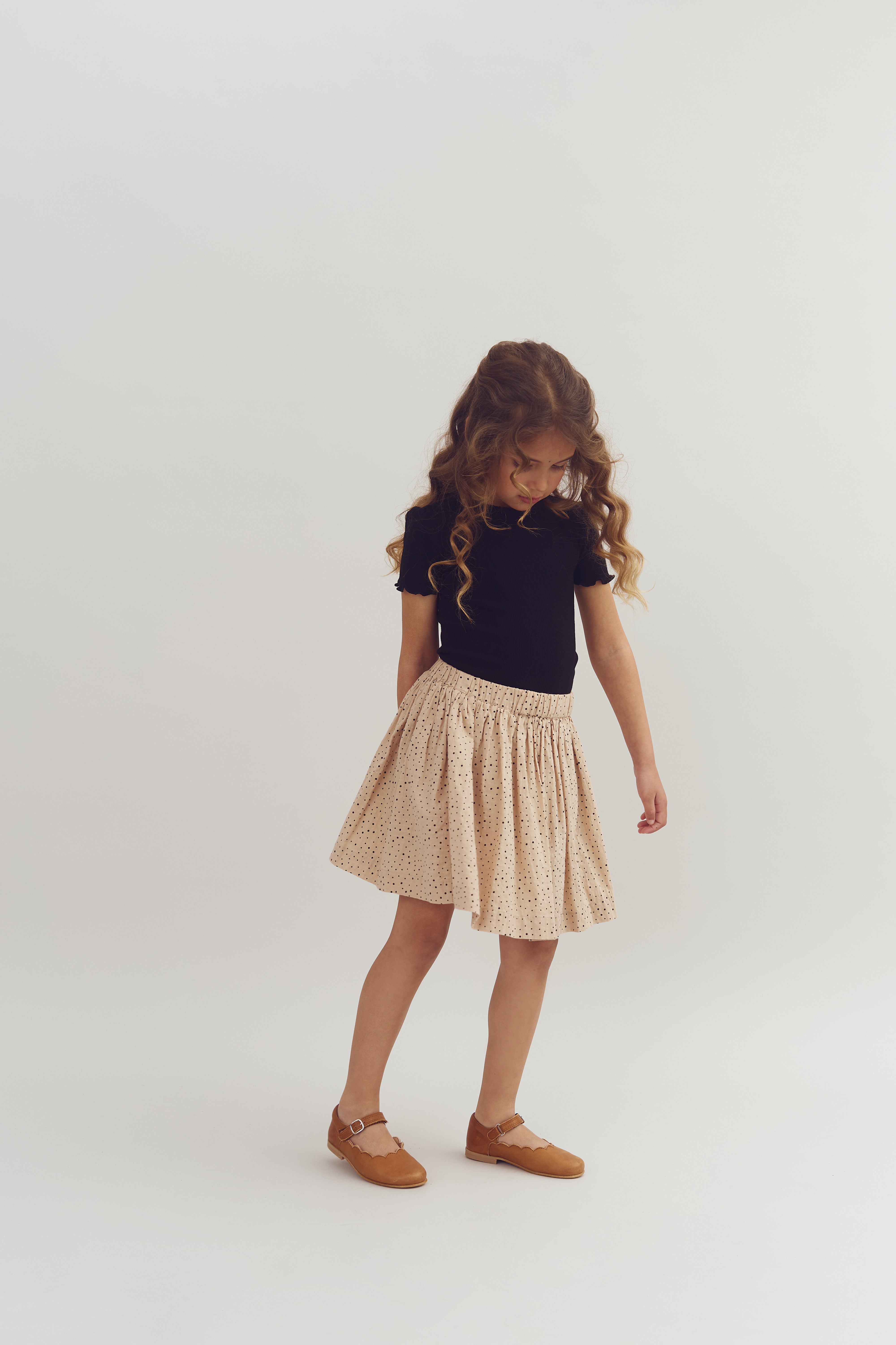 A young girl with long, curly hair stands looking down, wearing a black top, beige polka dot skirt, and brown shoes, set against a plain, light background.