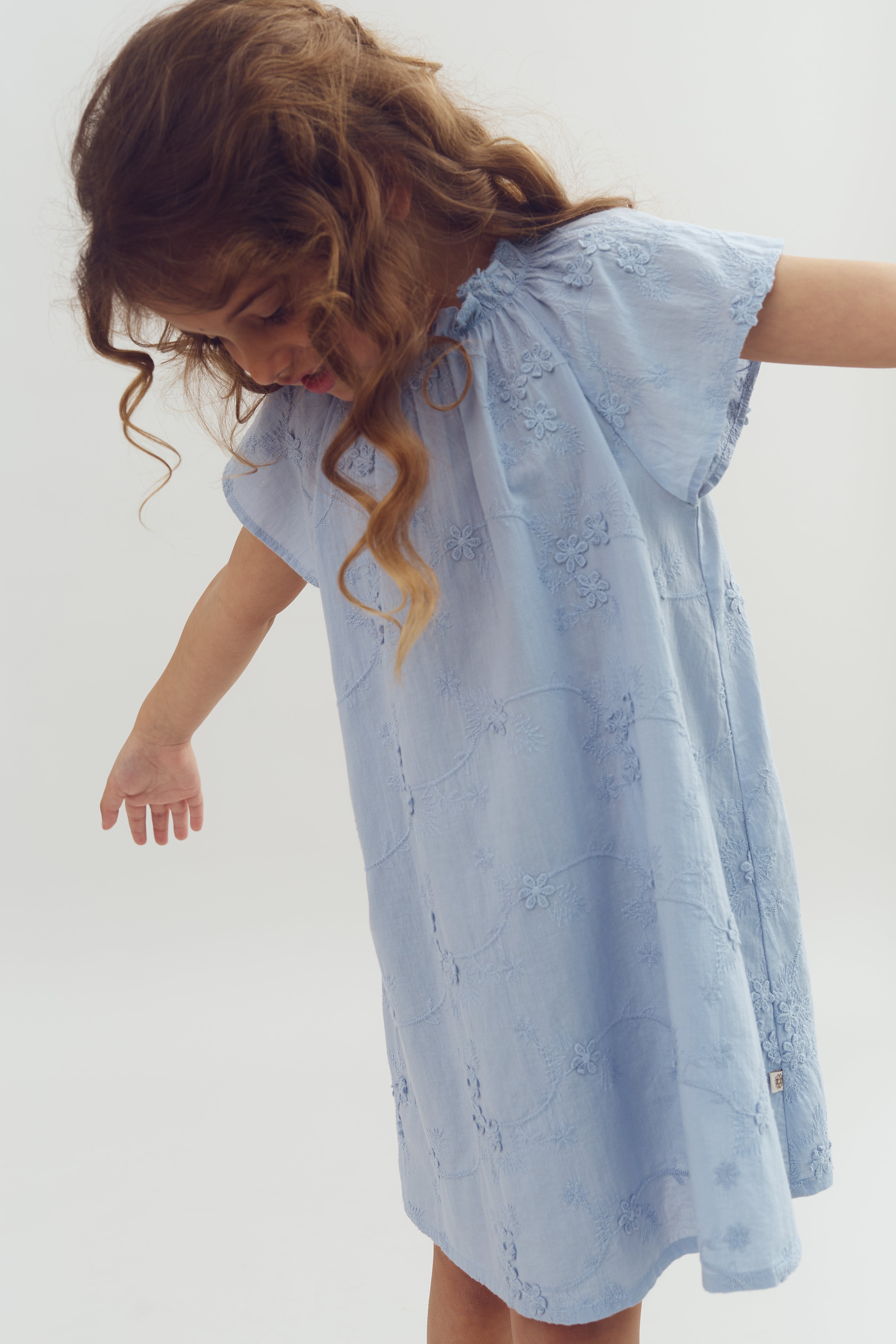 A young child wearing a light blue, embroidered dress gently leans forward, arms outstretched. The background is a plain white, emphasizing the child's playful movement.