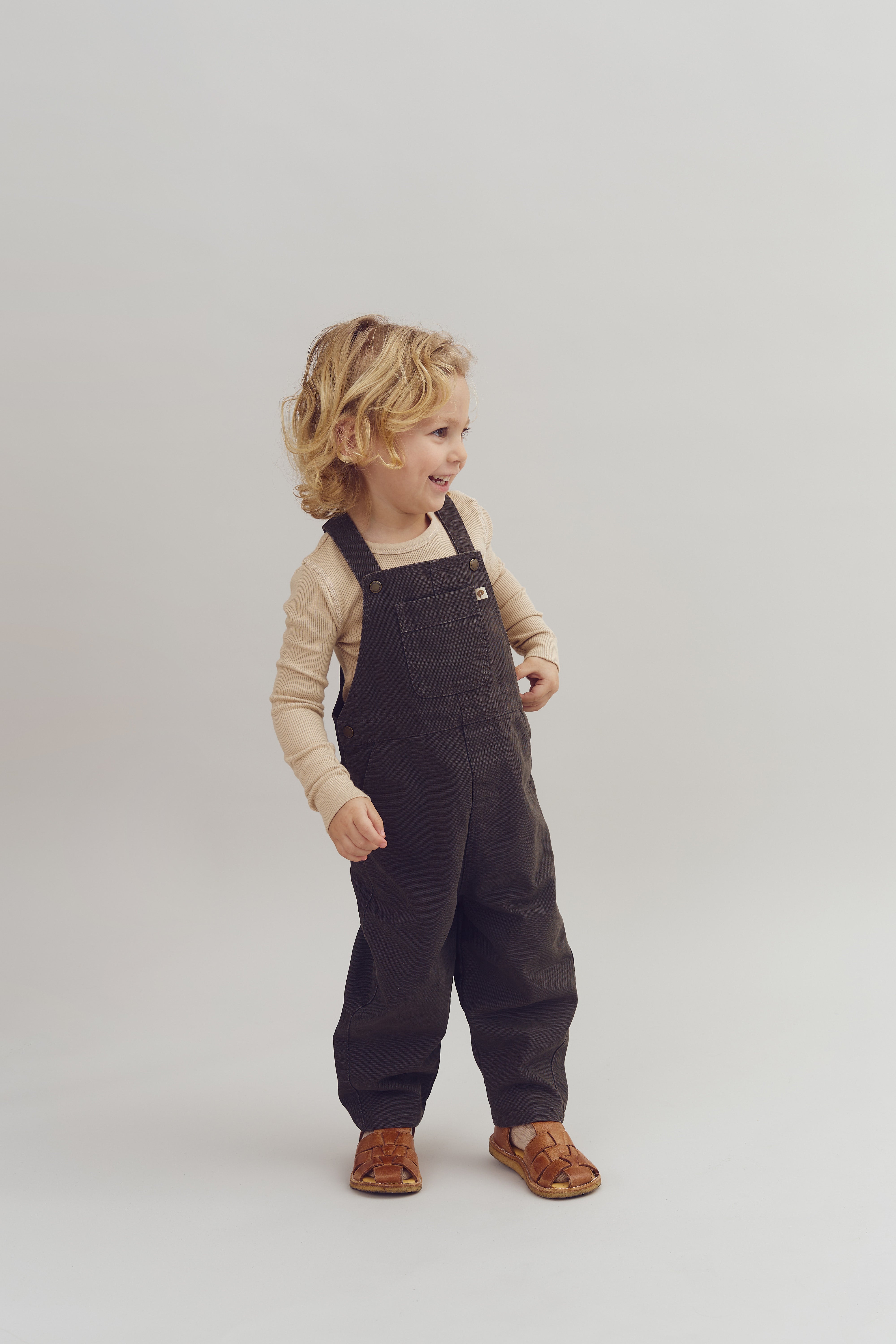 Barn iført en lysebrun trøje og mørke overalls smiler, mens det står på en lys baggrund. Det har krøllet, blondt hår og brune sko, og udstråler glæde.