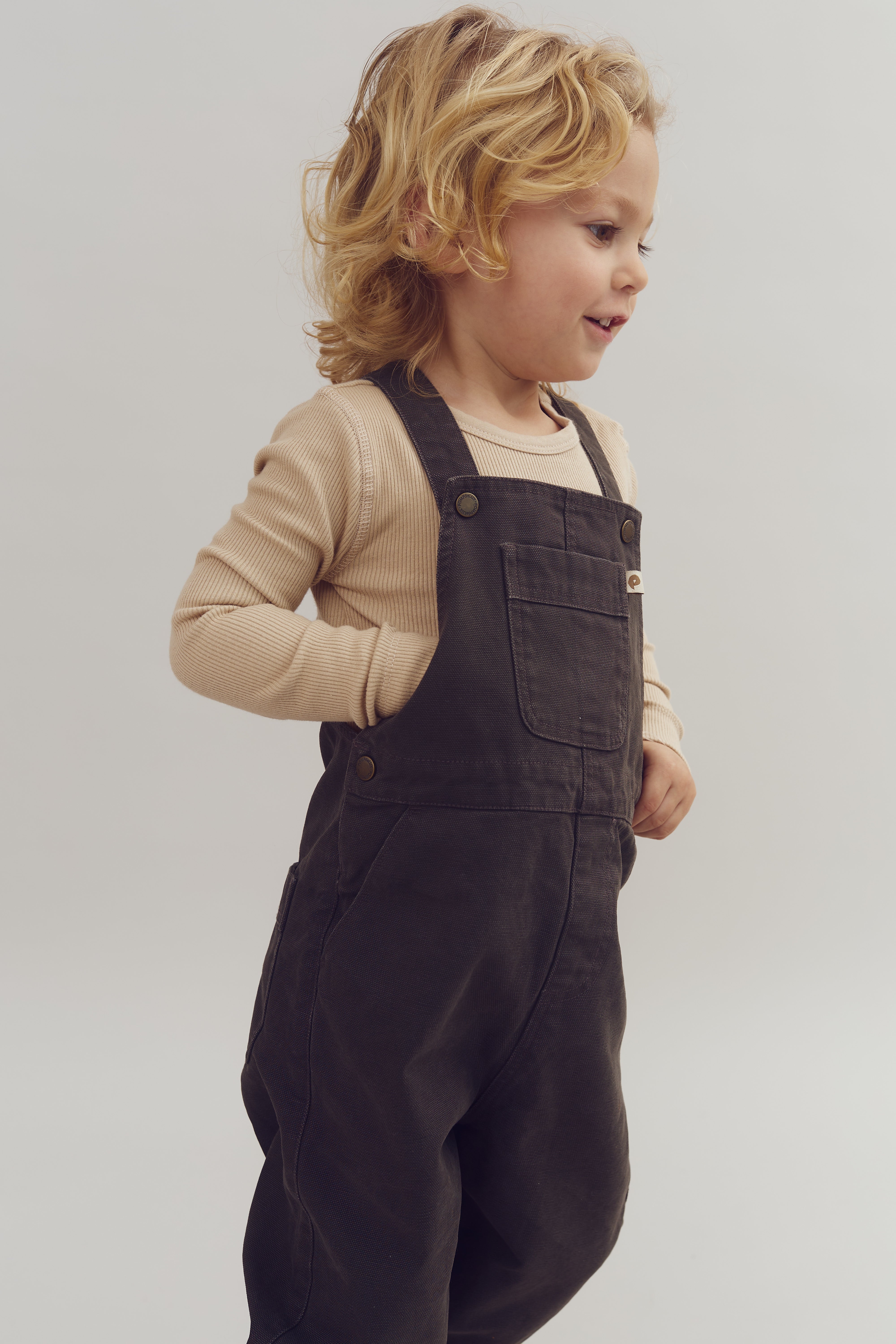 Et barn med lyst, krøllet hår bærer mørke overalls og en lysebrun trøje. Barnet smiler og står i en neutral baggrund med hænderne i lommerne.