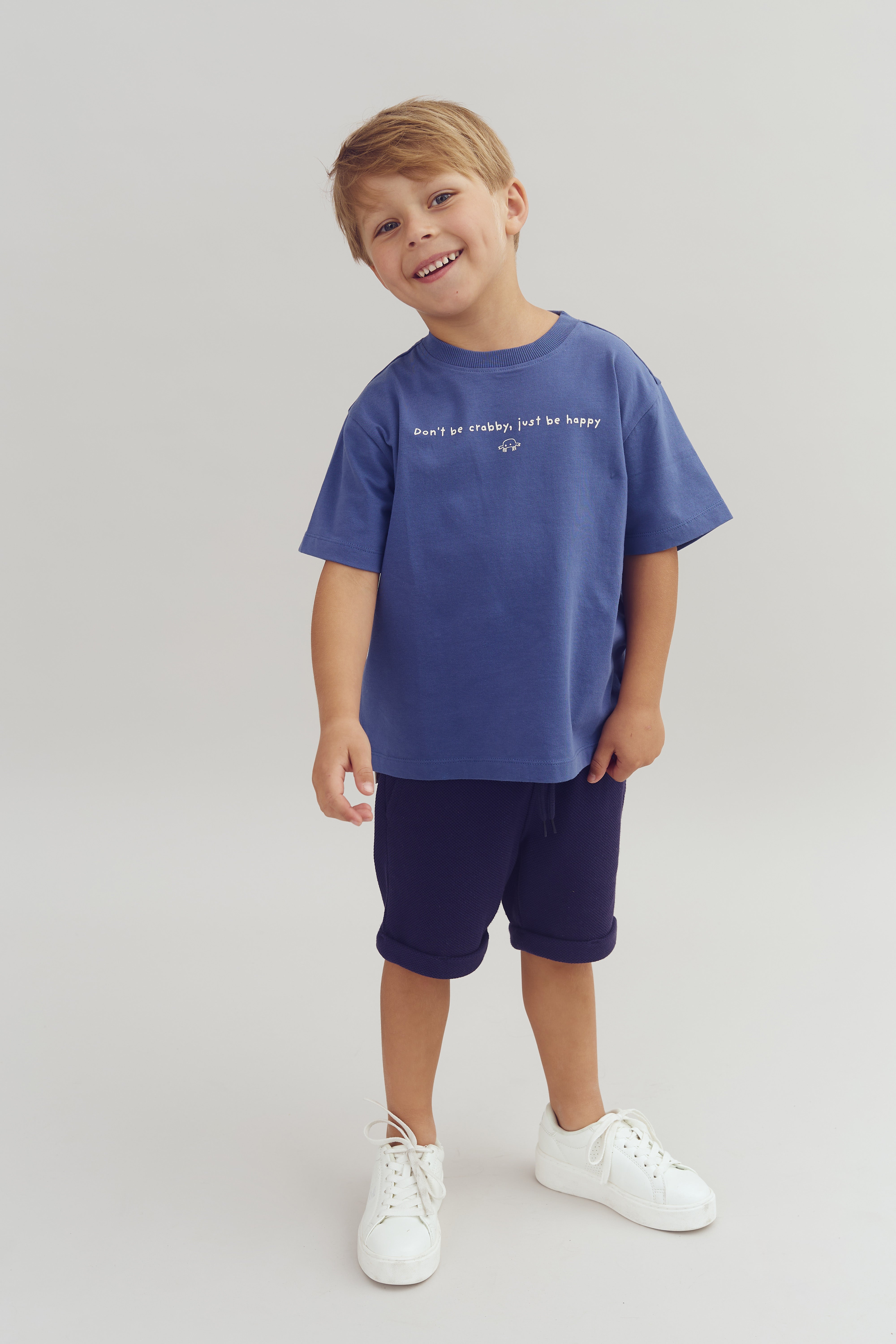 Barn står smilende iført blå t-shirt og shorts i en lys, neutral baggrund. Skjorten har teksten "Don't be crabby, just be happy." Han bærer hvide sneakers.