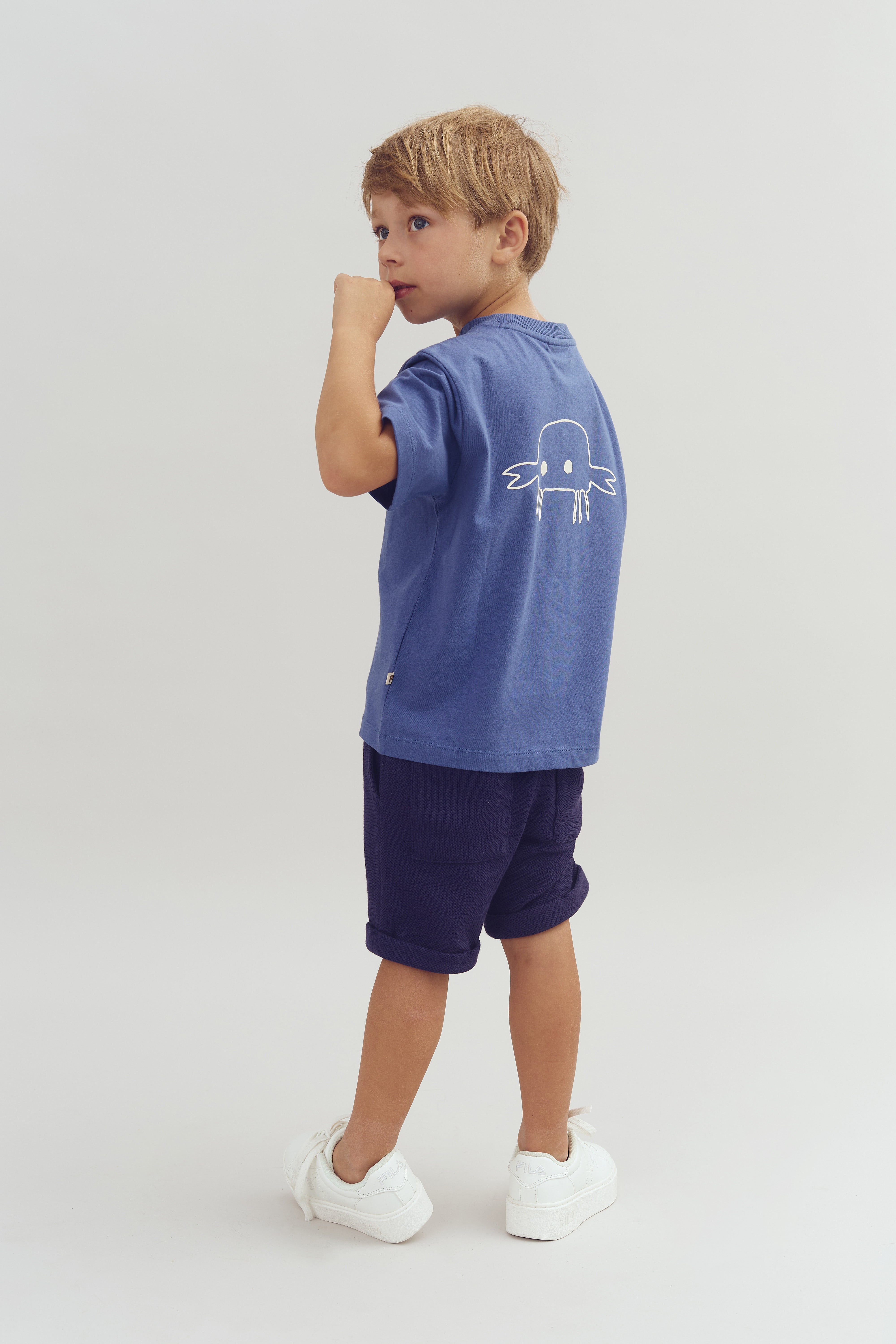 Dreng iført blå t-shirt med en hvid tegning på ryggen, står med hævet arm i en minimalistisk hvid baggrund, iført mørke shorts og hvide sko.