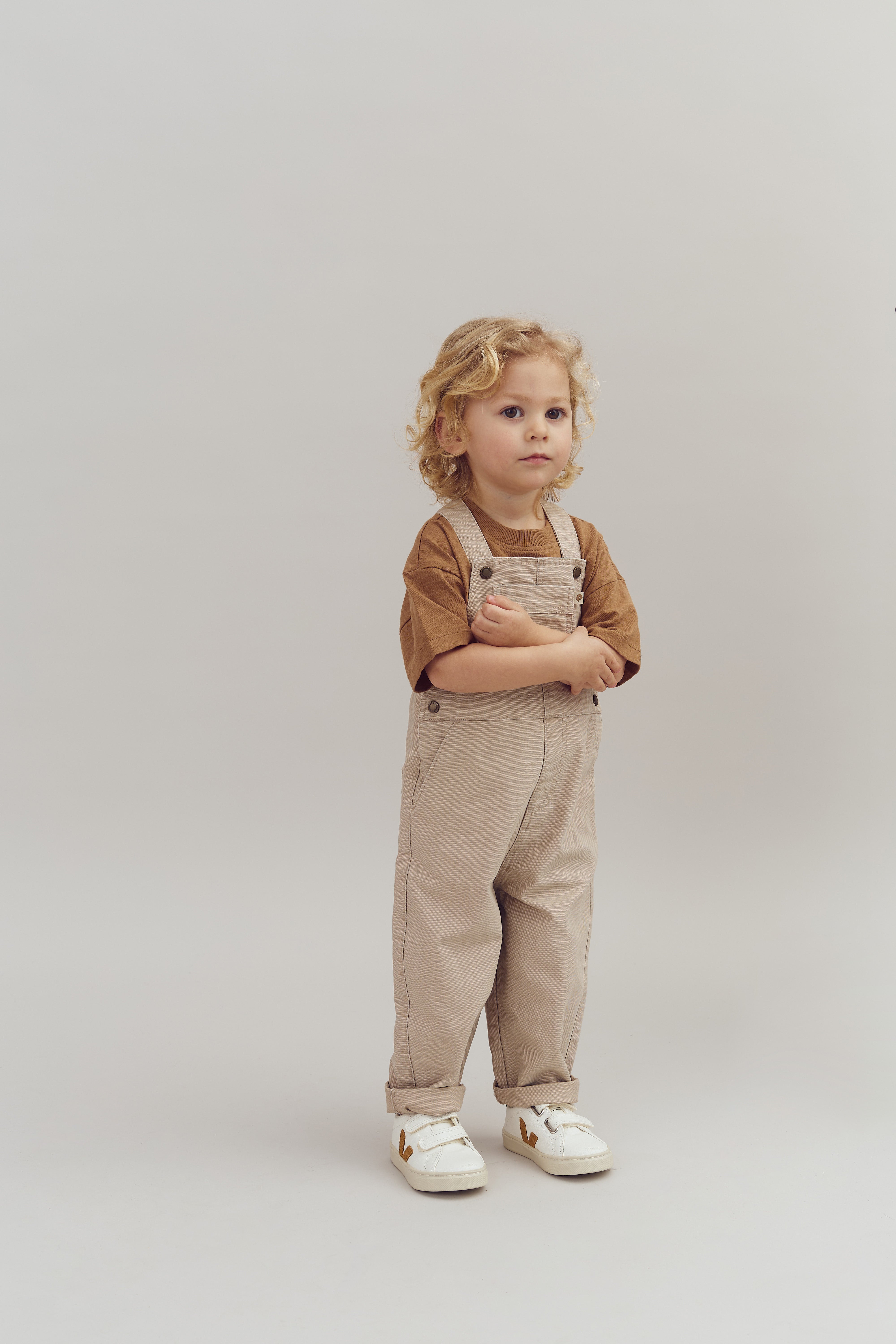 Et barn står afslappet i et par beige overalls med en brun t-shirt, omfavner sine arme i en enkel, grå baggrundsopsætning.