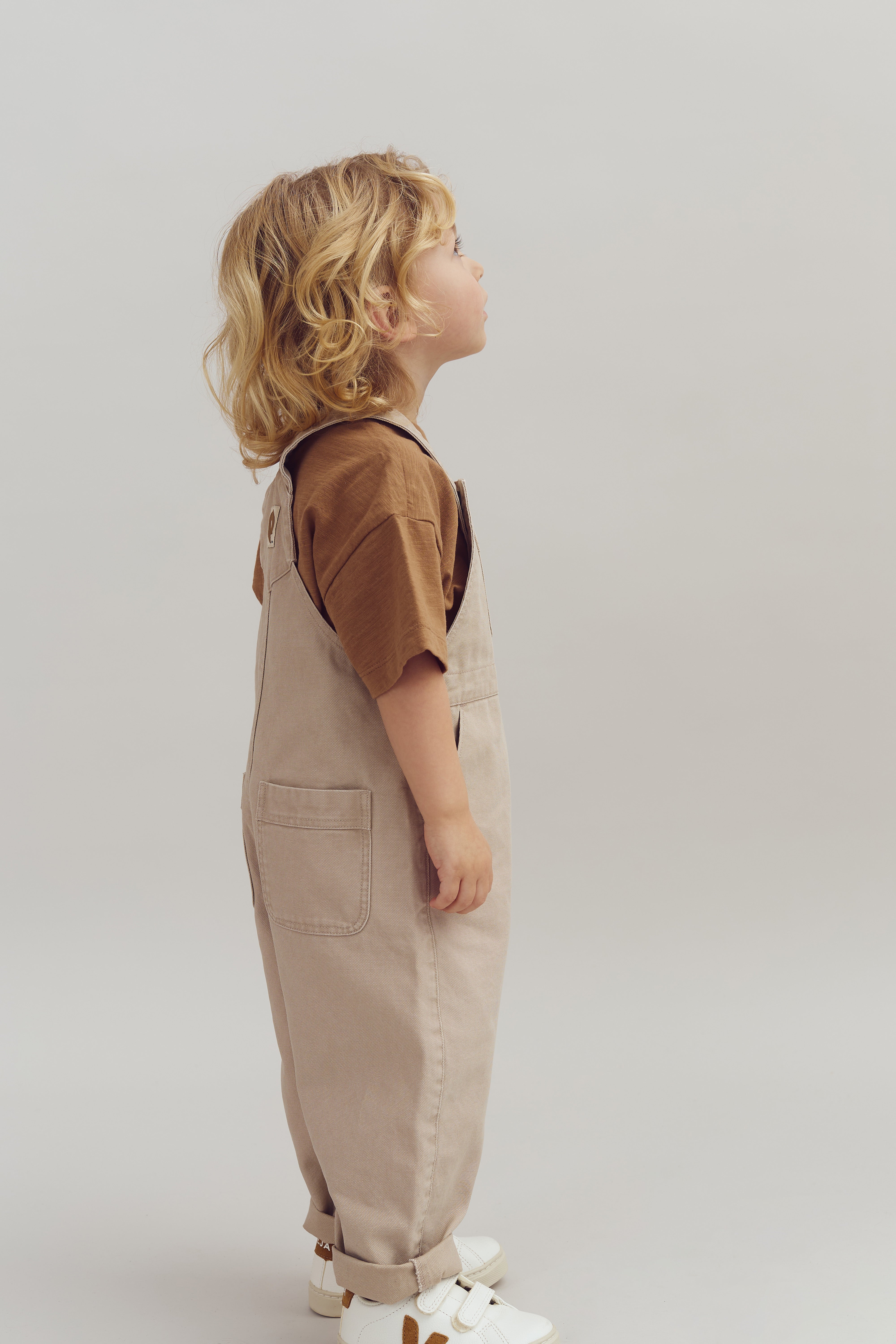 Barn står i profil, iført beige overalls og brun trøje, kigger opad, omgivet af en neutral, lysegrå baggrund.