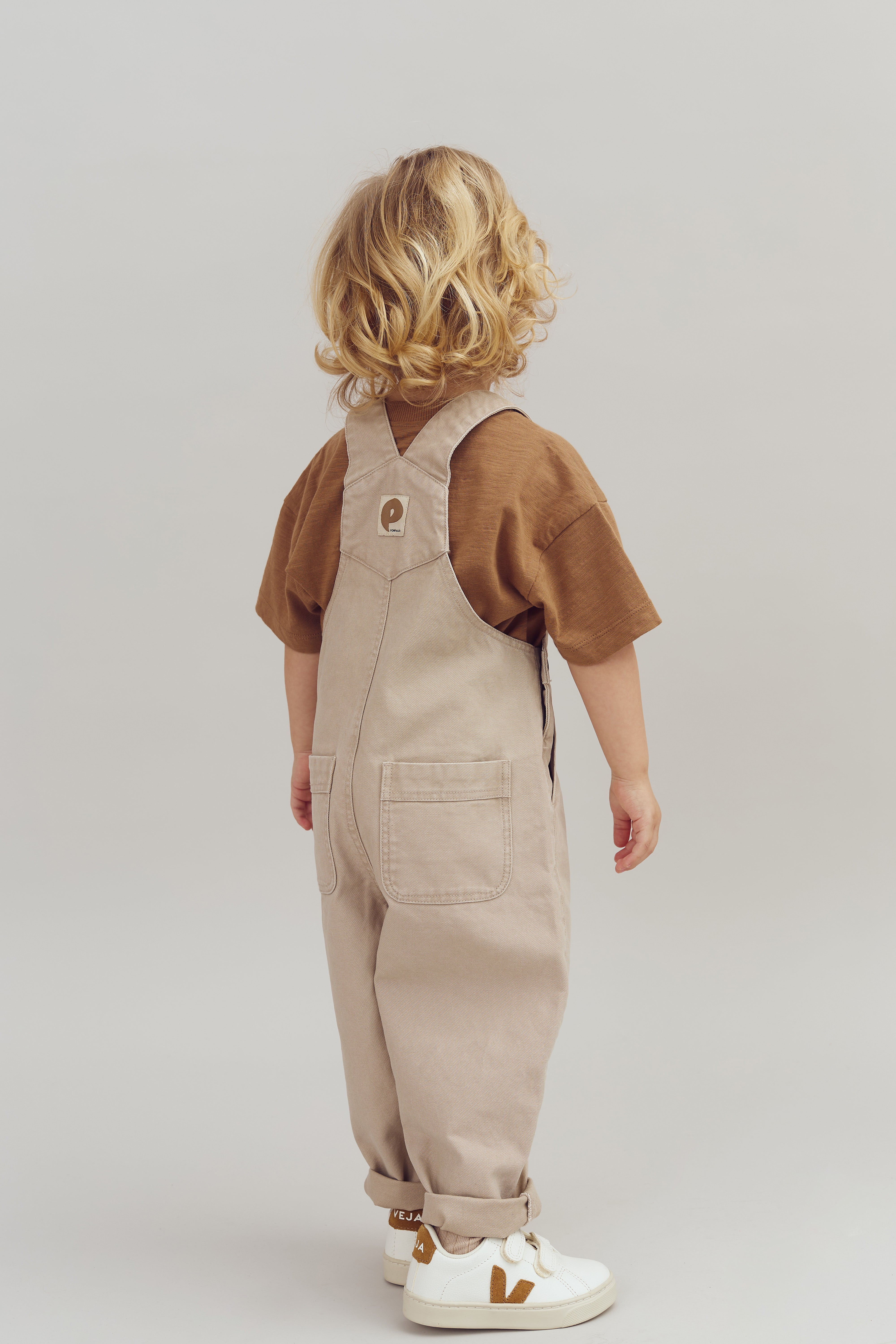 Et barn står med ryggen til iført beige overalls og brun trøje, i et neutralt, lyst rum. Barnets hår er blondt og krøllet. Sneakers har teksten "VEJA".
