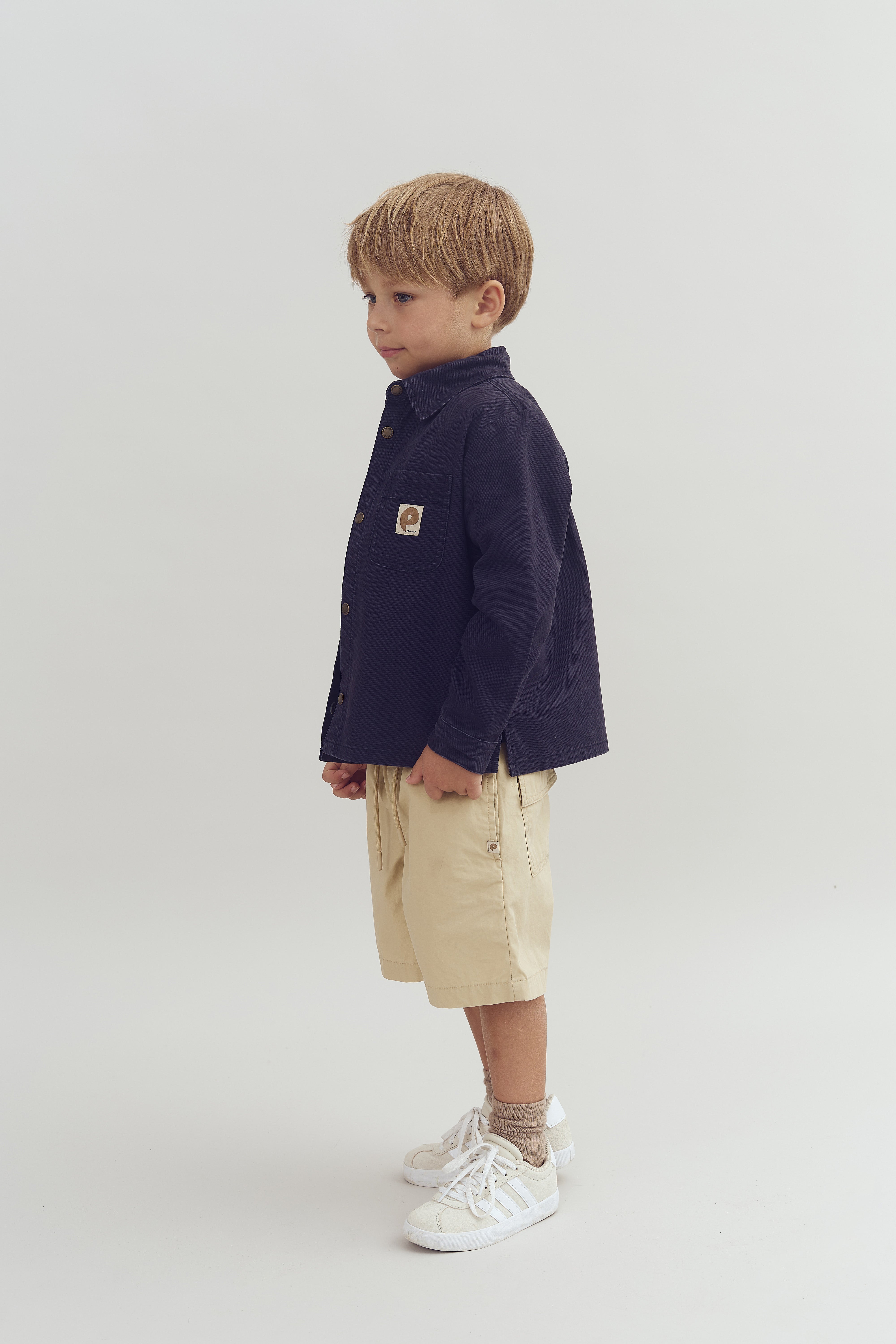Dreng står iført en mørk jakke med et mærke på lommen, beige shorts og hvide sneakers. Baggrunden er enkel og lys, hvilket fremhæver drengen.