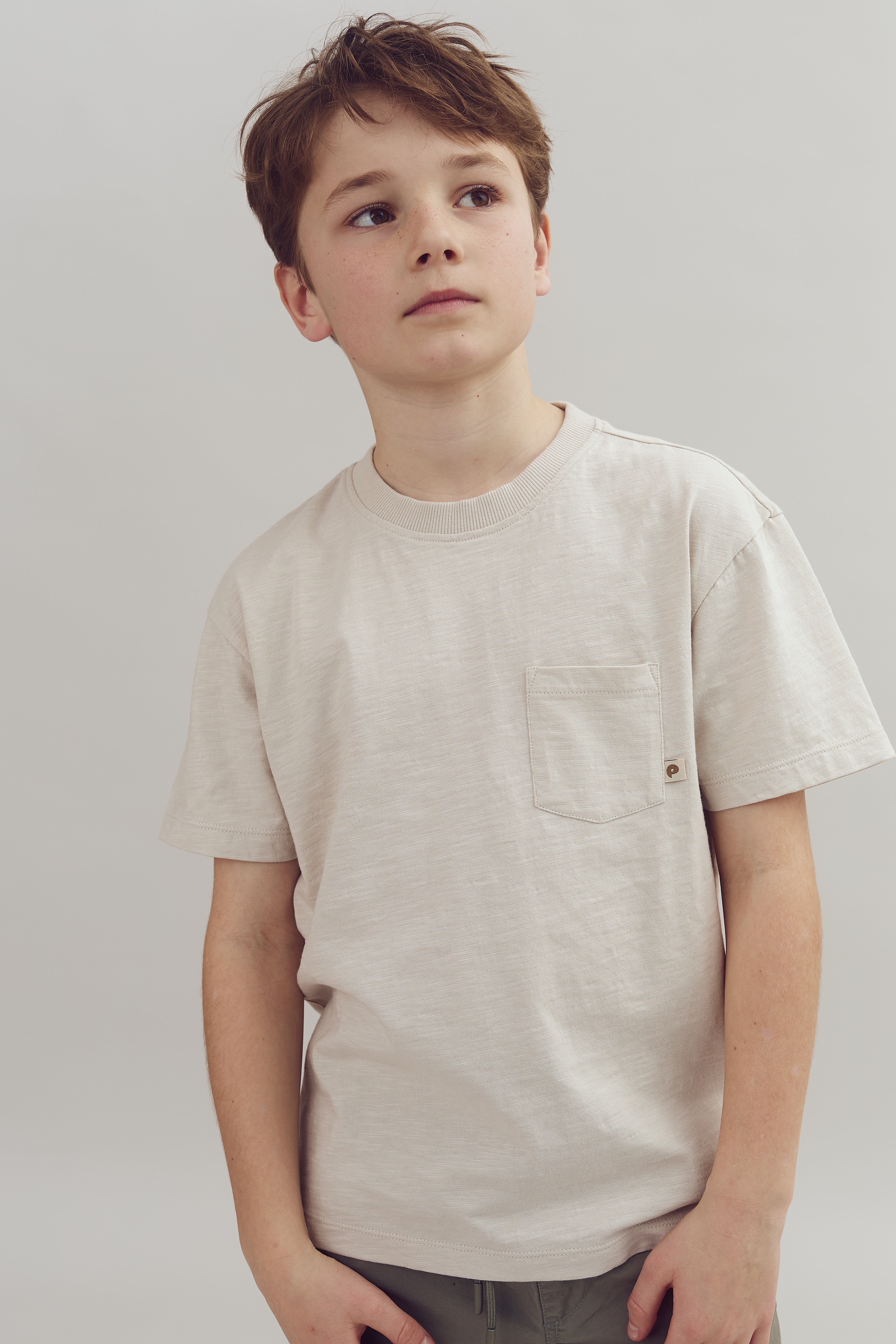 Dreng står iført en lys T-shirt med brystlomme, ser opad. Baggrunden er neutral og ensfarvet, hvilket fremhæver drengen som hovedfokus.