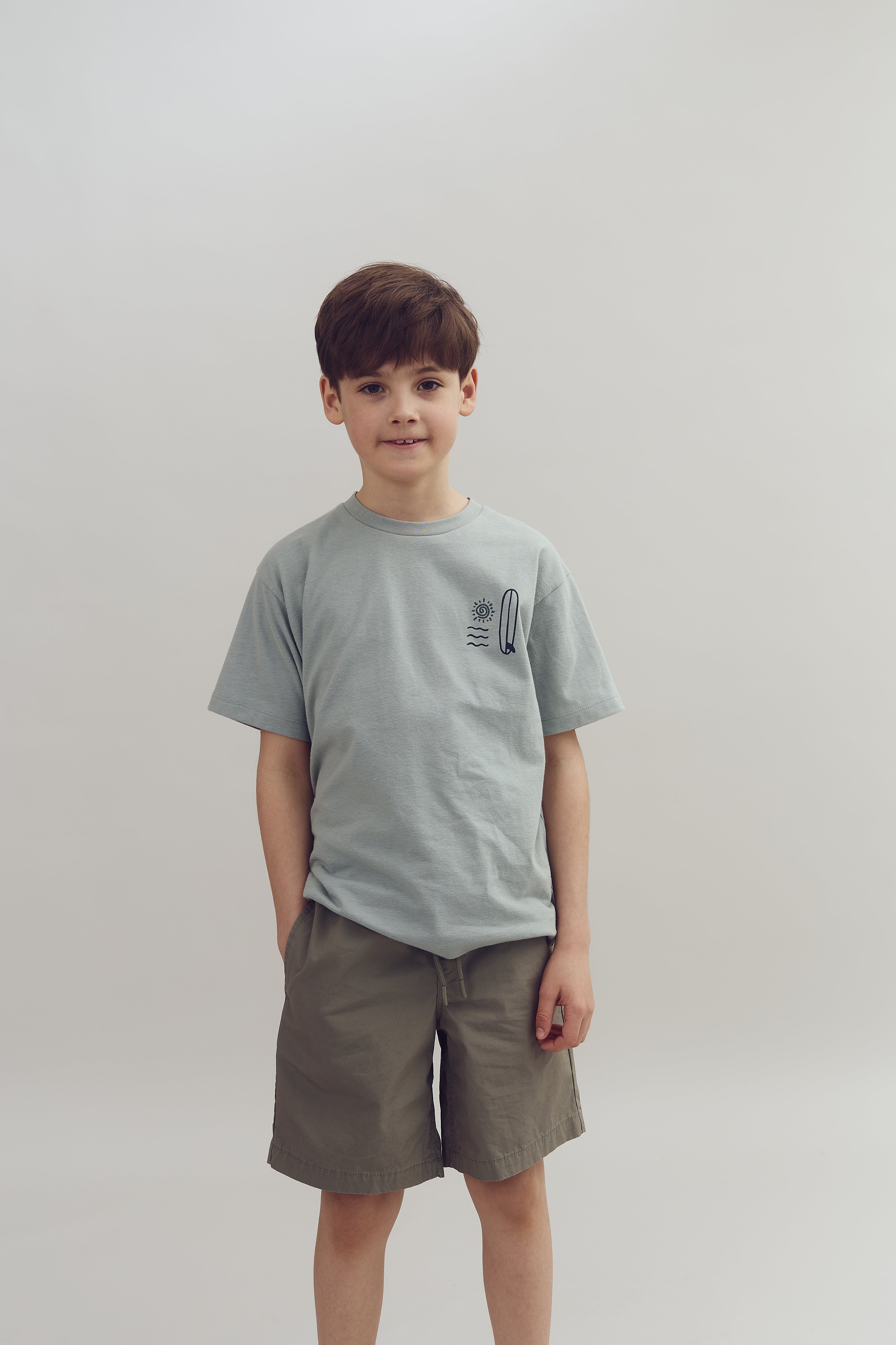 Dreng står stille iført en lysegrå T-shirt og grønne shorts i en neutral, lys baggrund. Trøjen har et tryk af en sol, bølger og en surfbræt.