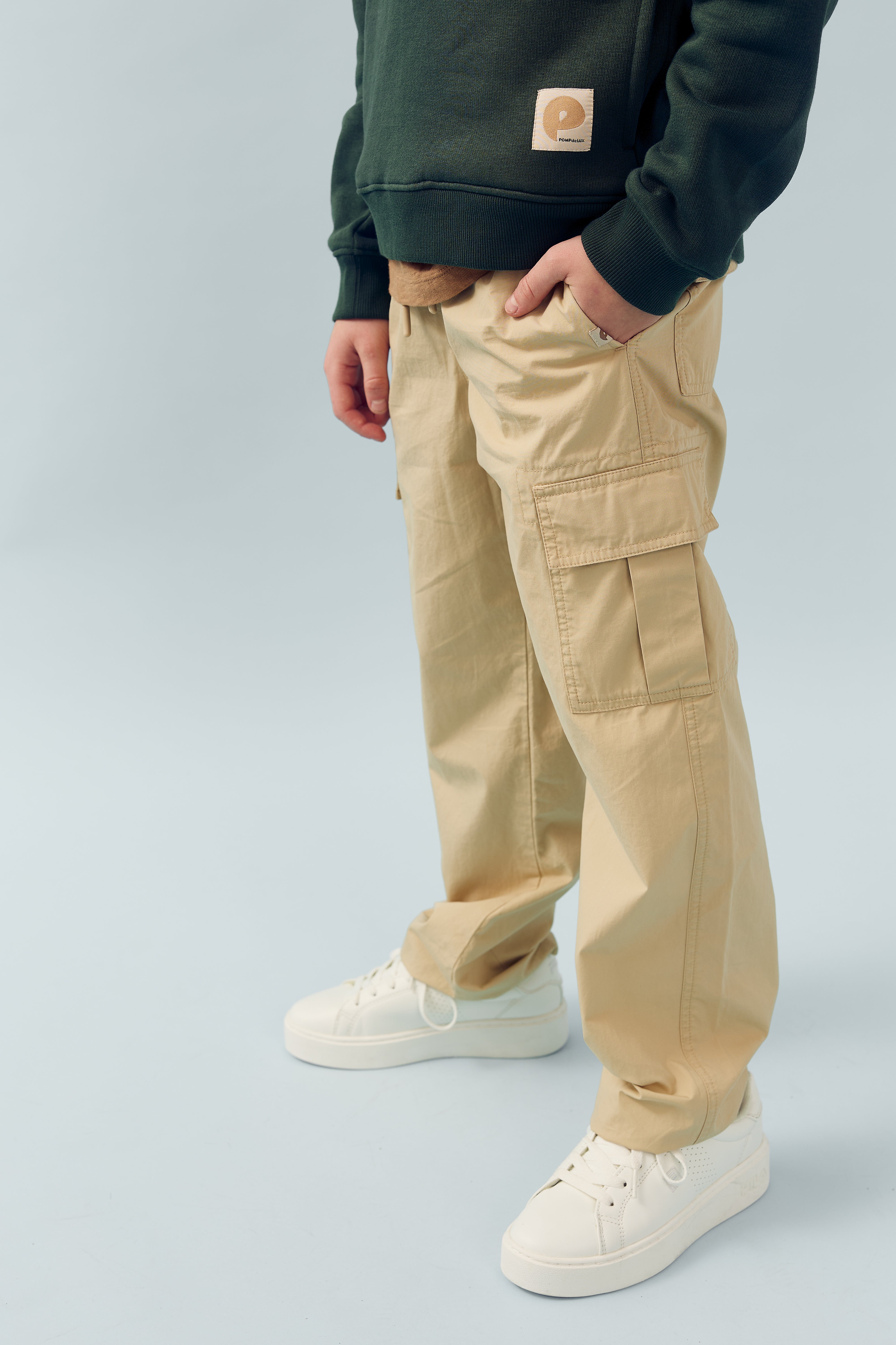 En person står iført beige cargo-bukser, grøn trøje med logo på lommen og hvide sneakers mod en lysblå baggrund. Personens venstre hånd er i lommen.
