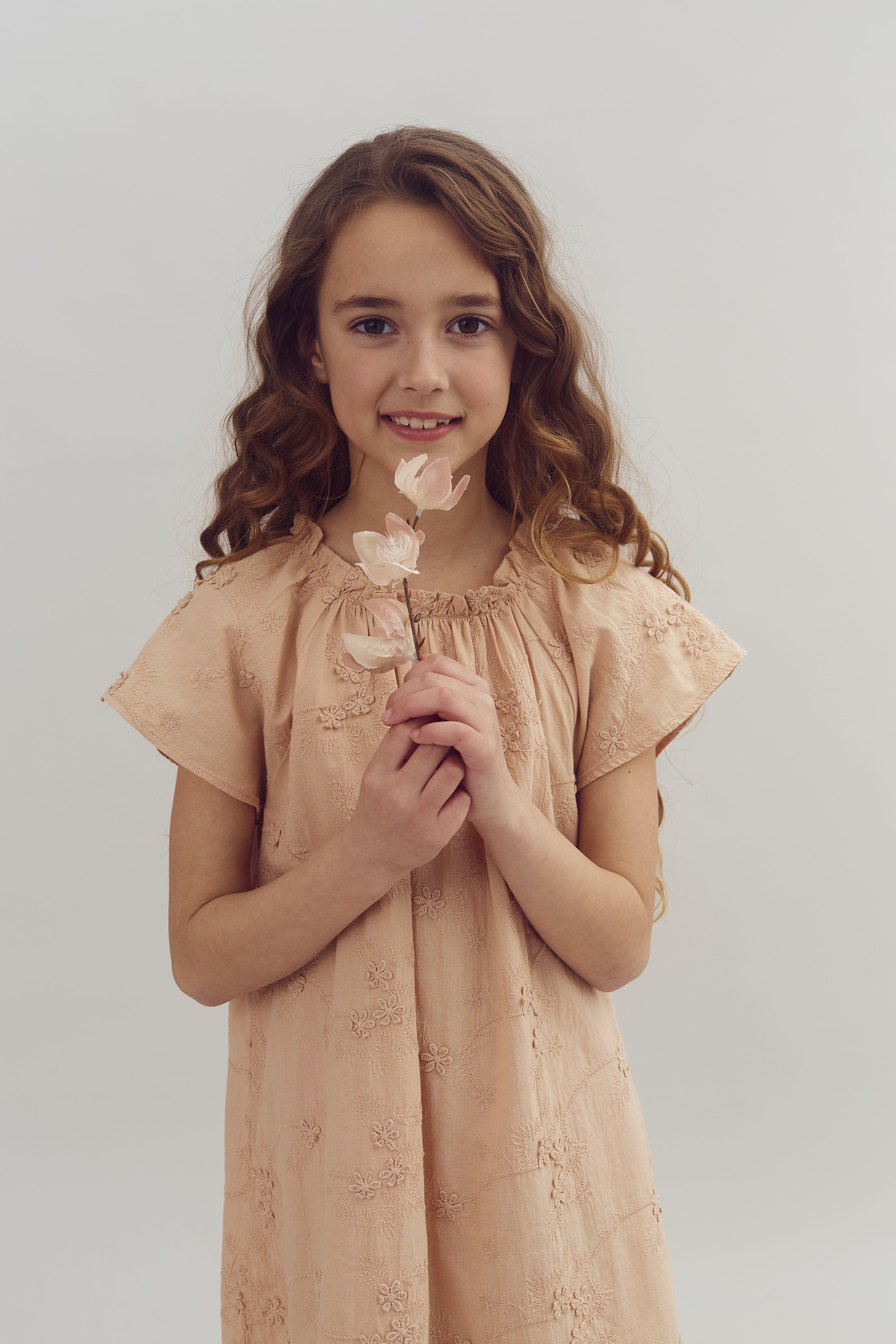 En pige holder en lille blomst, mens hun smiler. Hun bærer en lyserød kjole med broderede blomster. Baggrunden er enkel og lys.