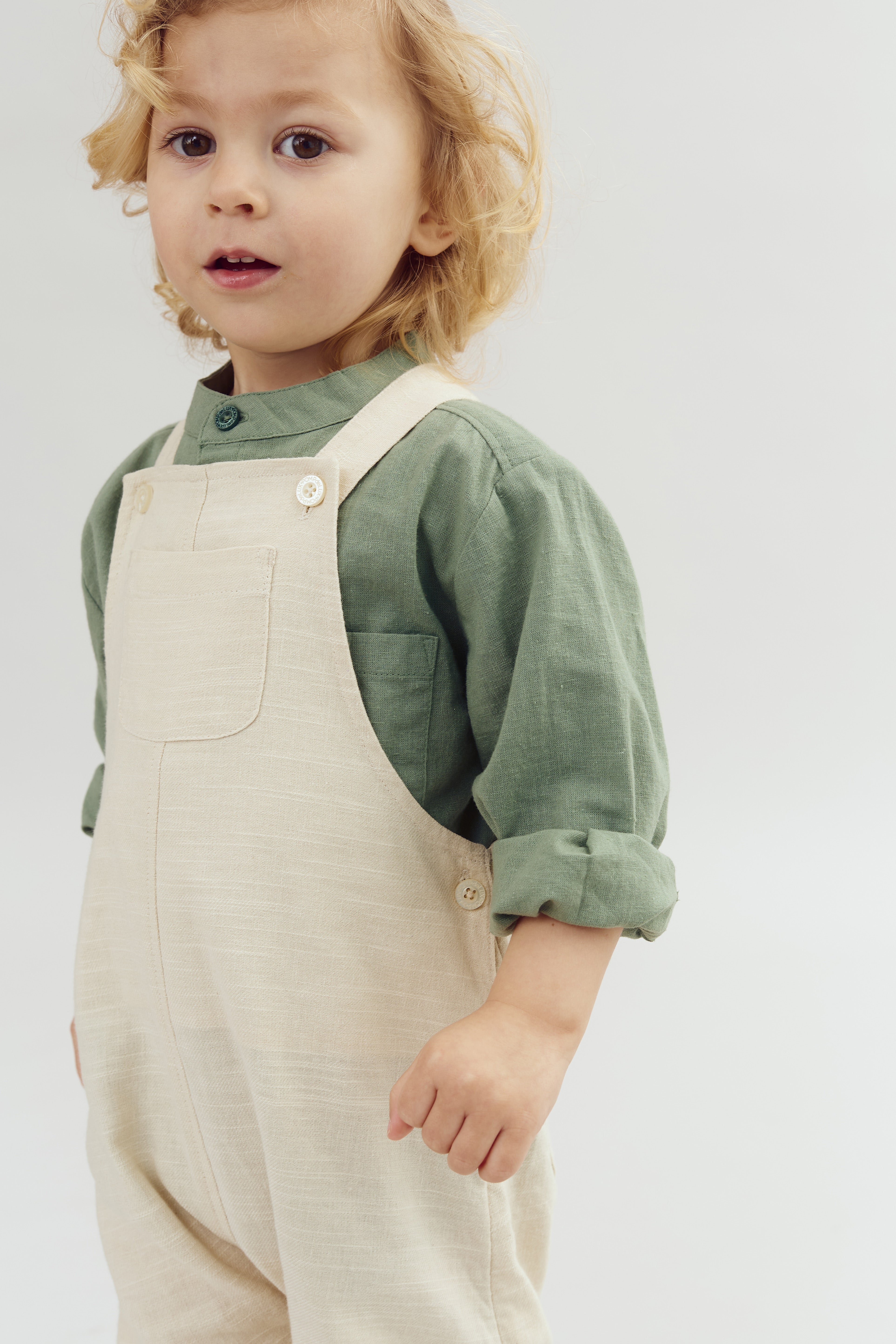 Barn i beige overalls og grøn skjorte står i neutrale omgivelser og kigger fremad med lys baggrund.
