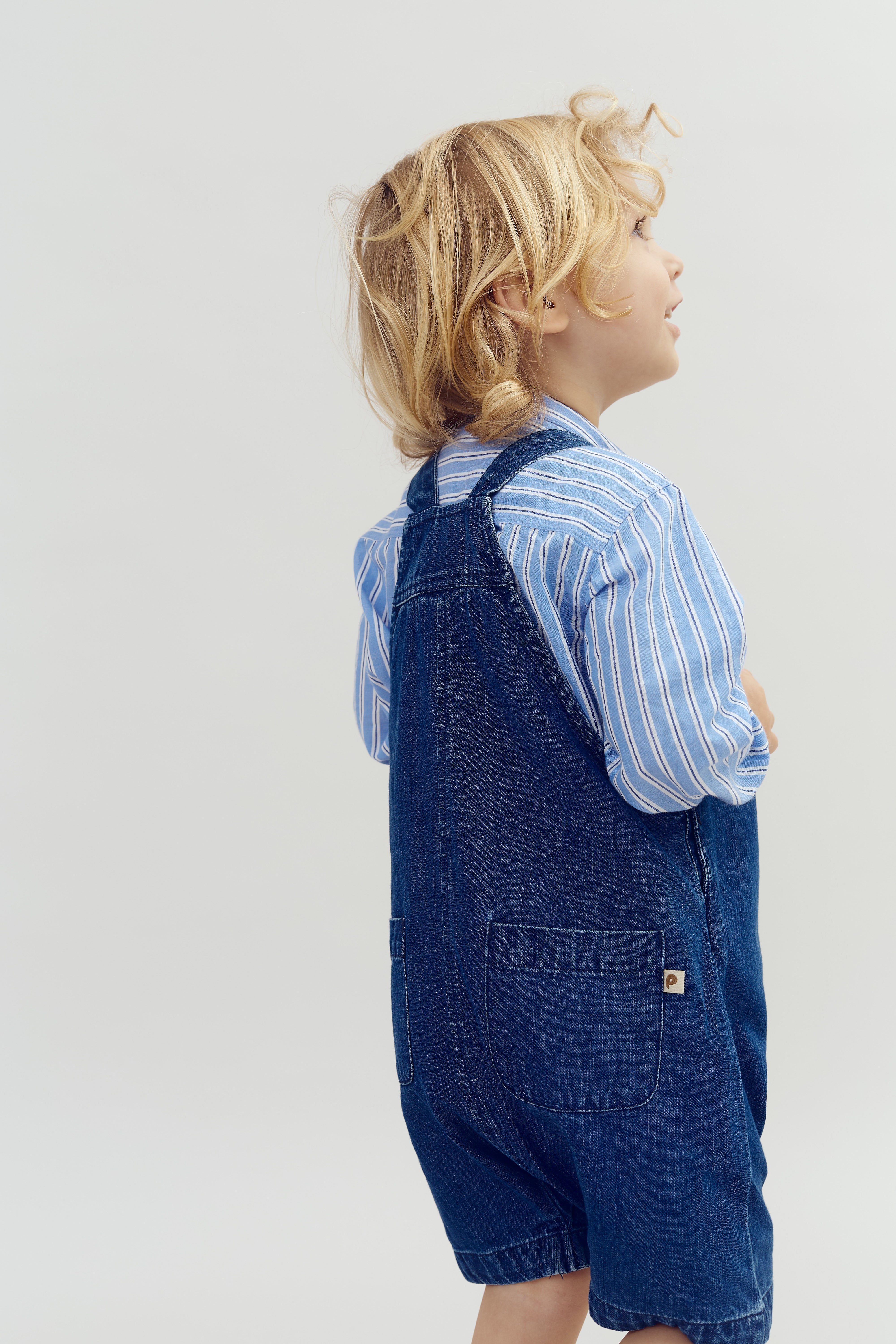 Et barn med blondt hår ser op, iført en blåstribet skjorte og denimshorts, mod en neutral baggrund.