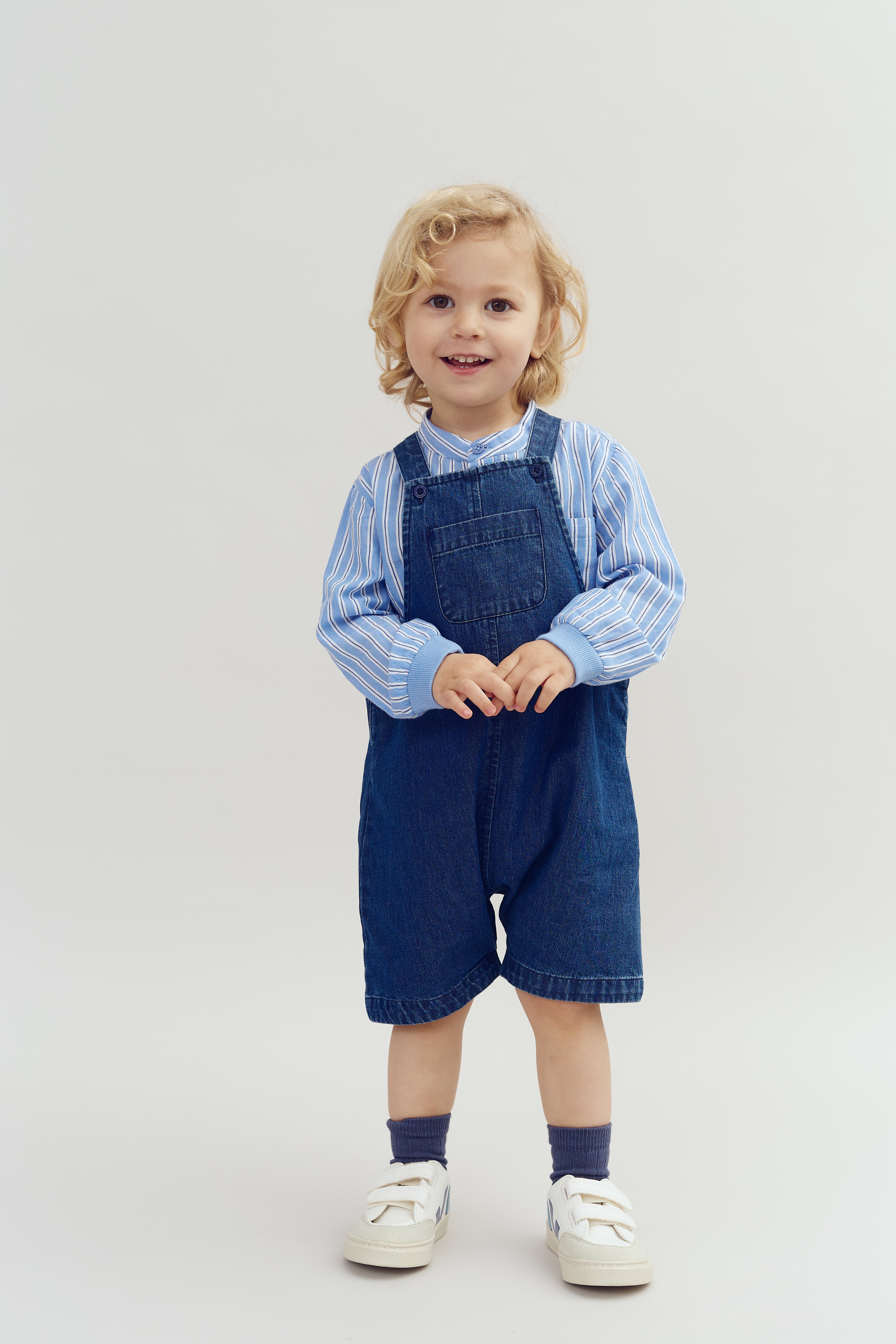 Barn står smilende iført blåstribet skjorte og denim-overalls, neutrale lyse baggrund.