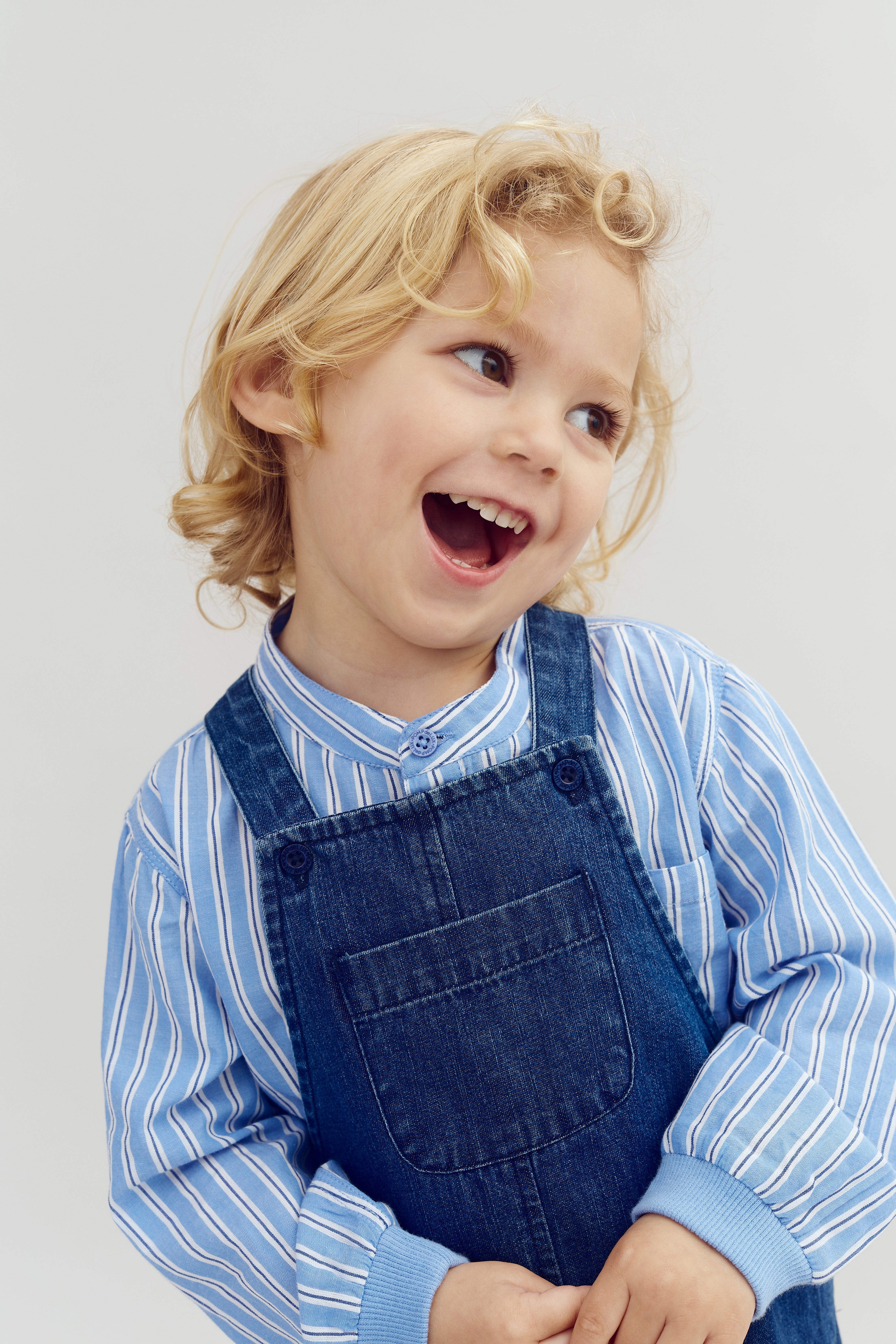 Child smiling joyfully, wearing denim overalls over a blue striped shirt. The background is plain and light-colored, emphasizing the child's cheerful expression.