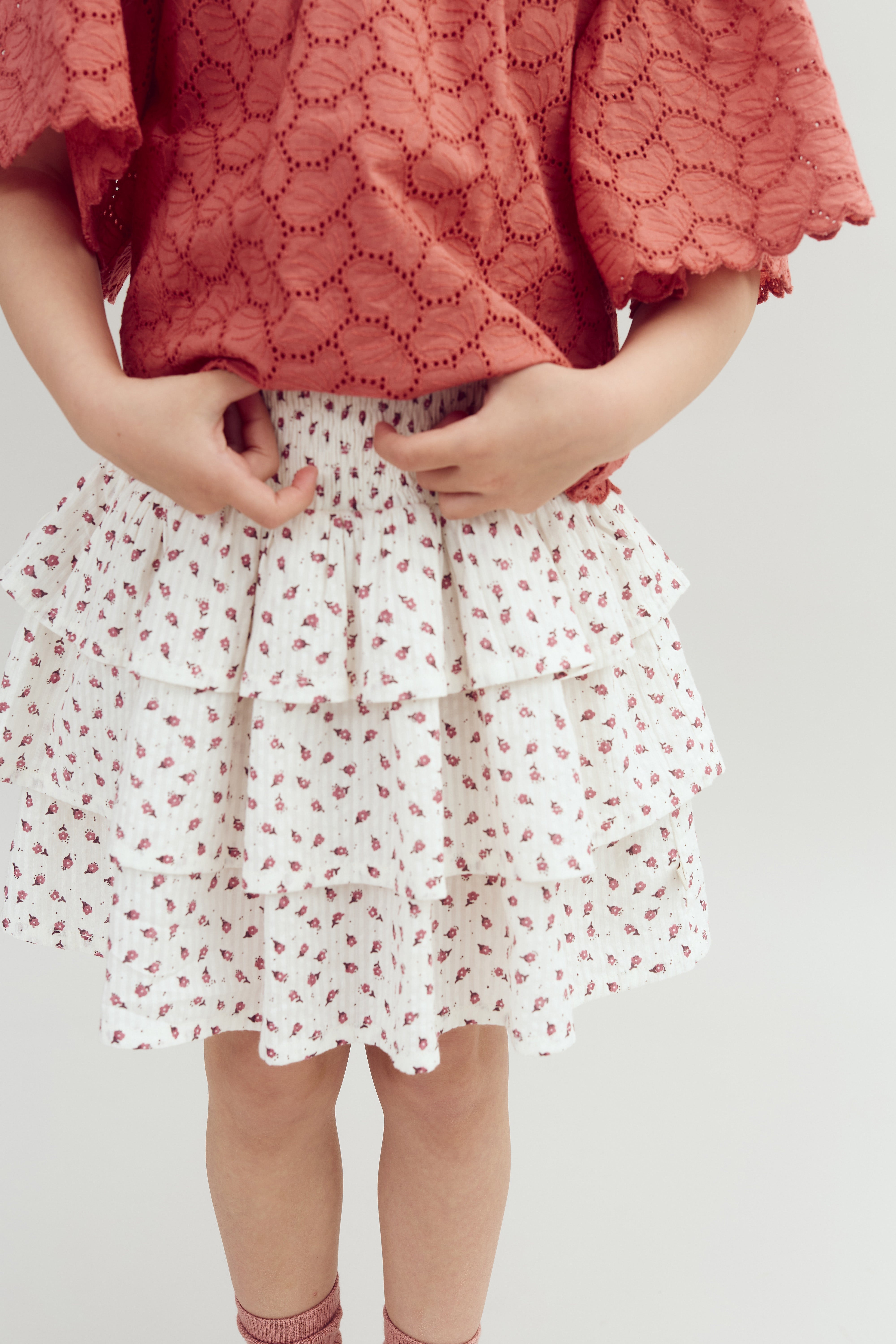 Child wearing a red lace top adjusts a white, tiered skirt with small floral patterns. The child’s hands are on the waistband in a neutral background.