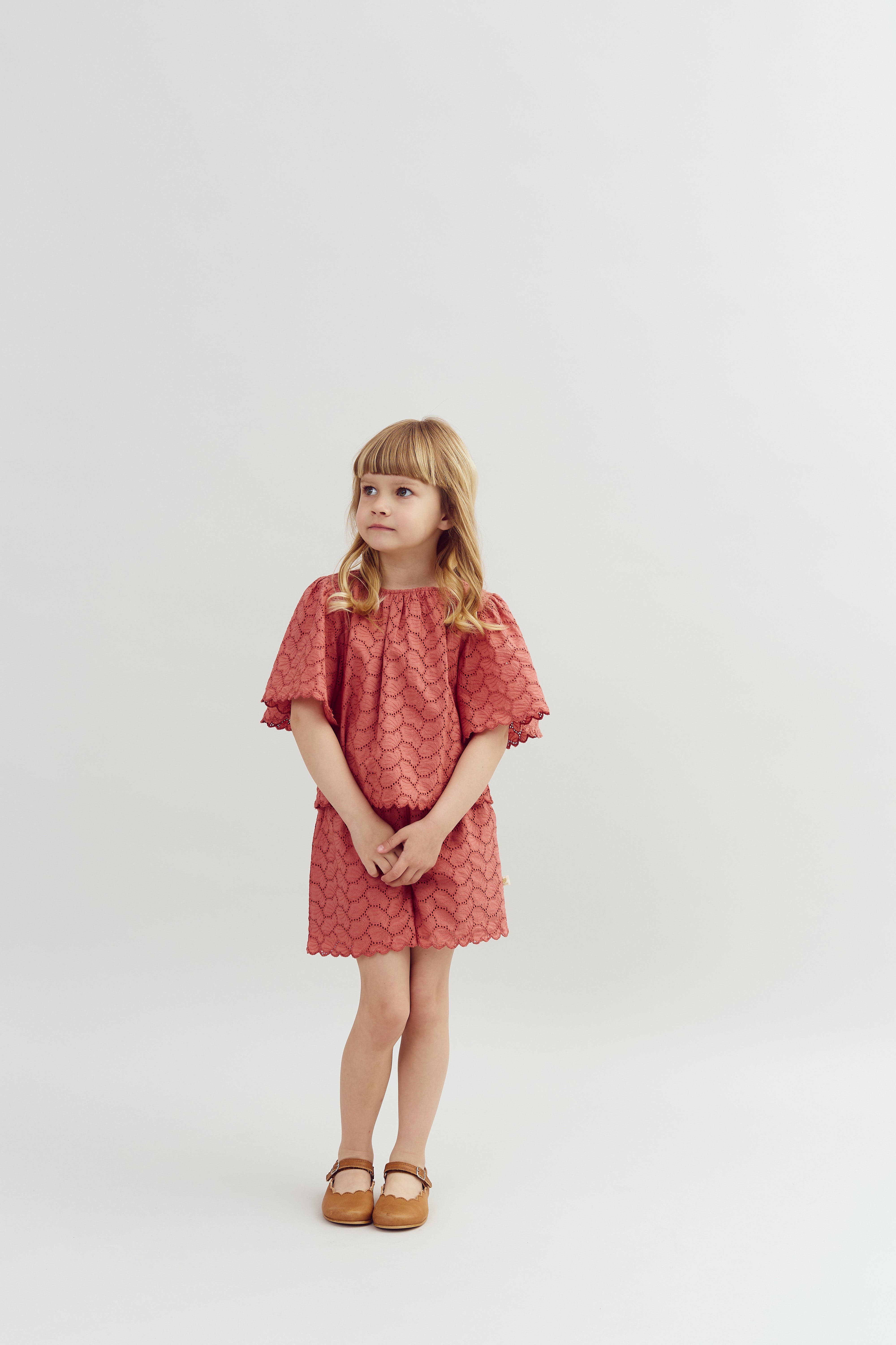 A young child stands, wearing a patterned pink dress and brown shoes, gazing upward in a plain, softly lit studio setting.