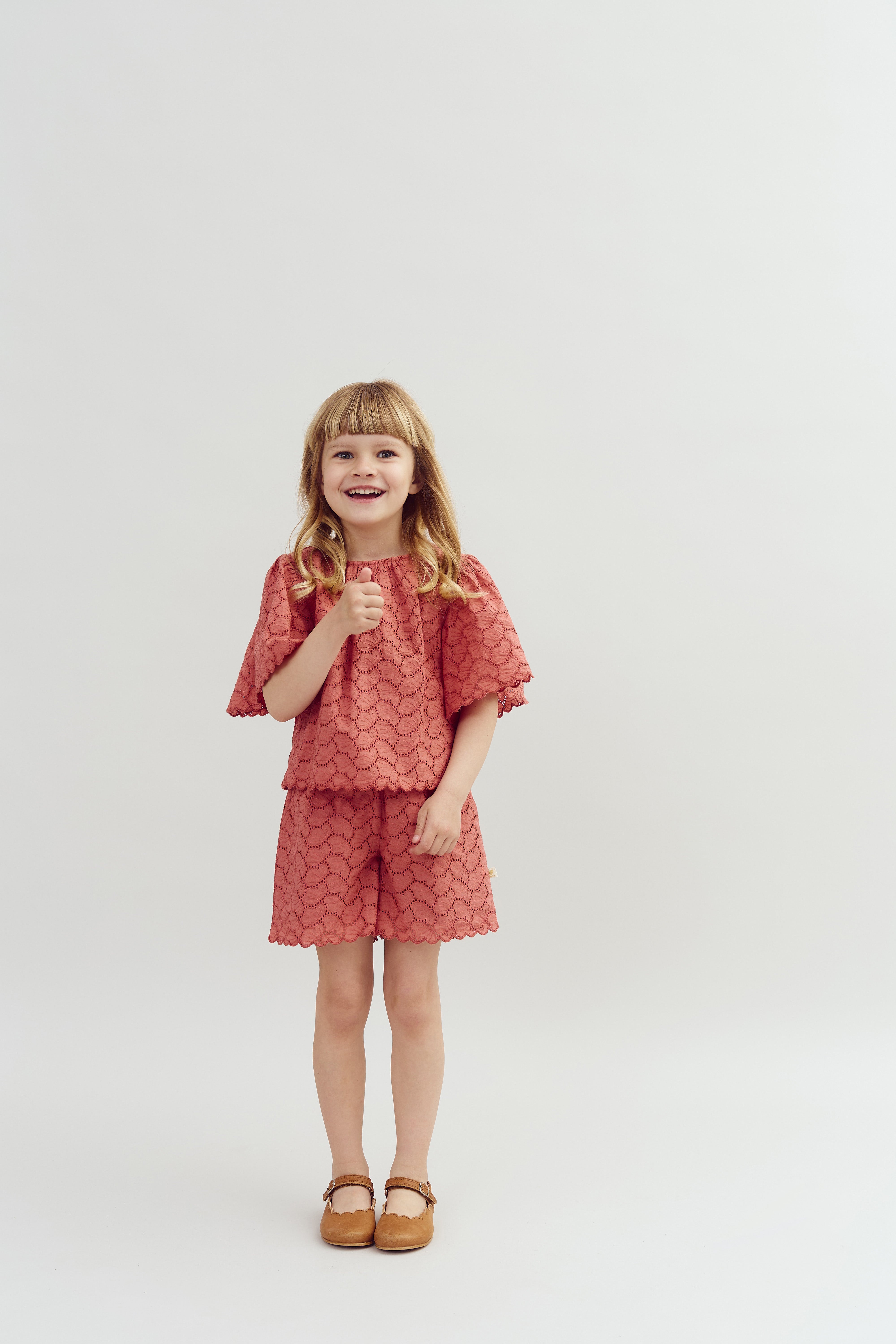 A young child stands smiling, giving a thumbs-up gesture, wearing a lace-patterned coral outfit and brown shoes, set against a plain white background.