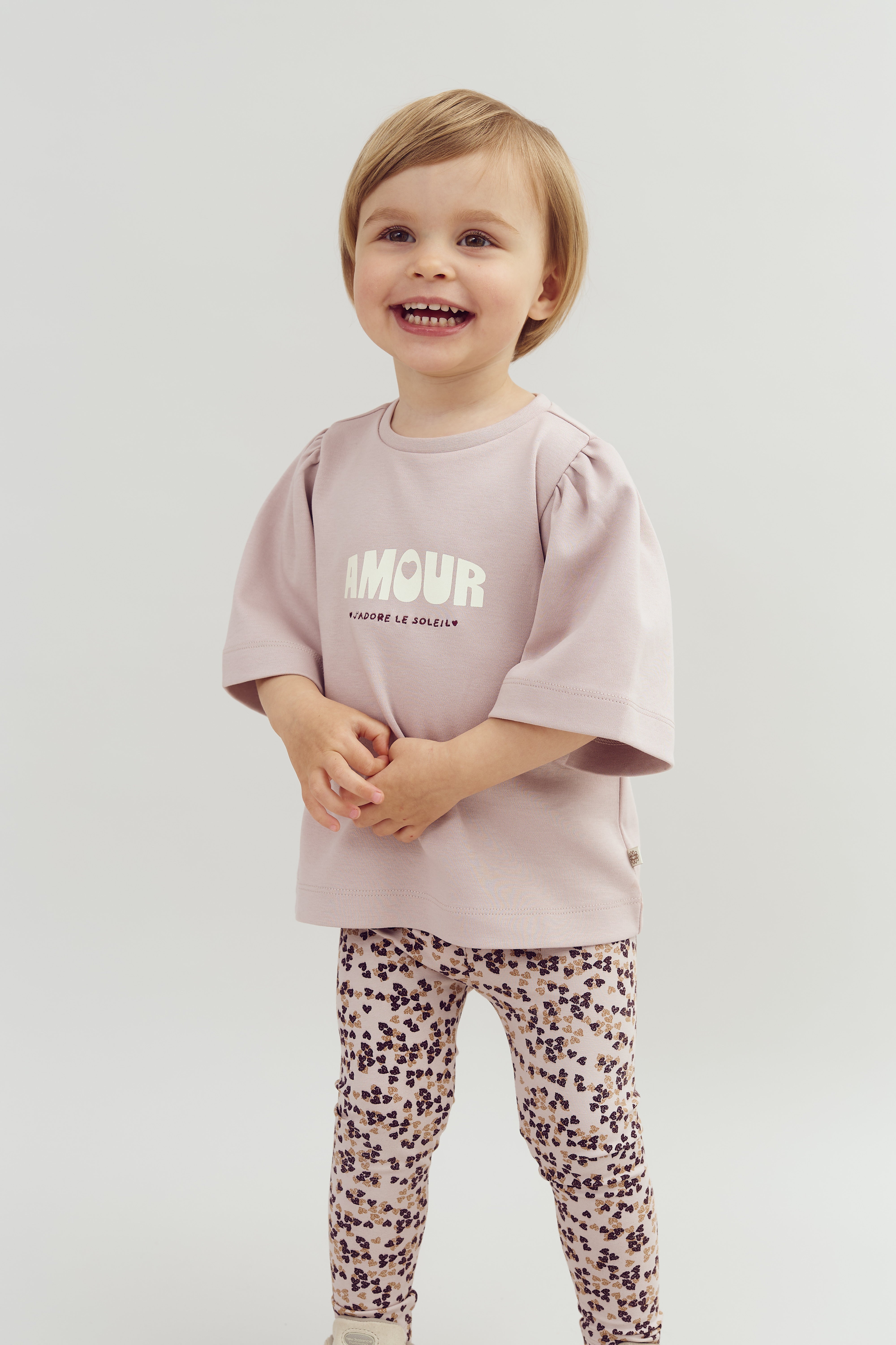Et barn smiler, iført en lys lilla trøje med teksten "AMOUR J'ADORE LE SOLEIL" og mønstrede leggings, stående mod en neutral baggrund.
