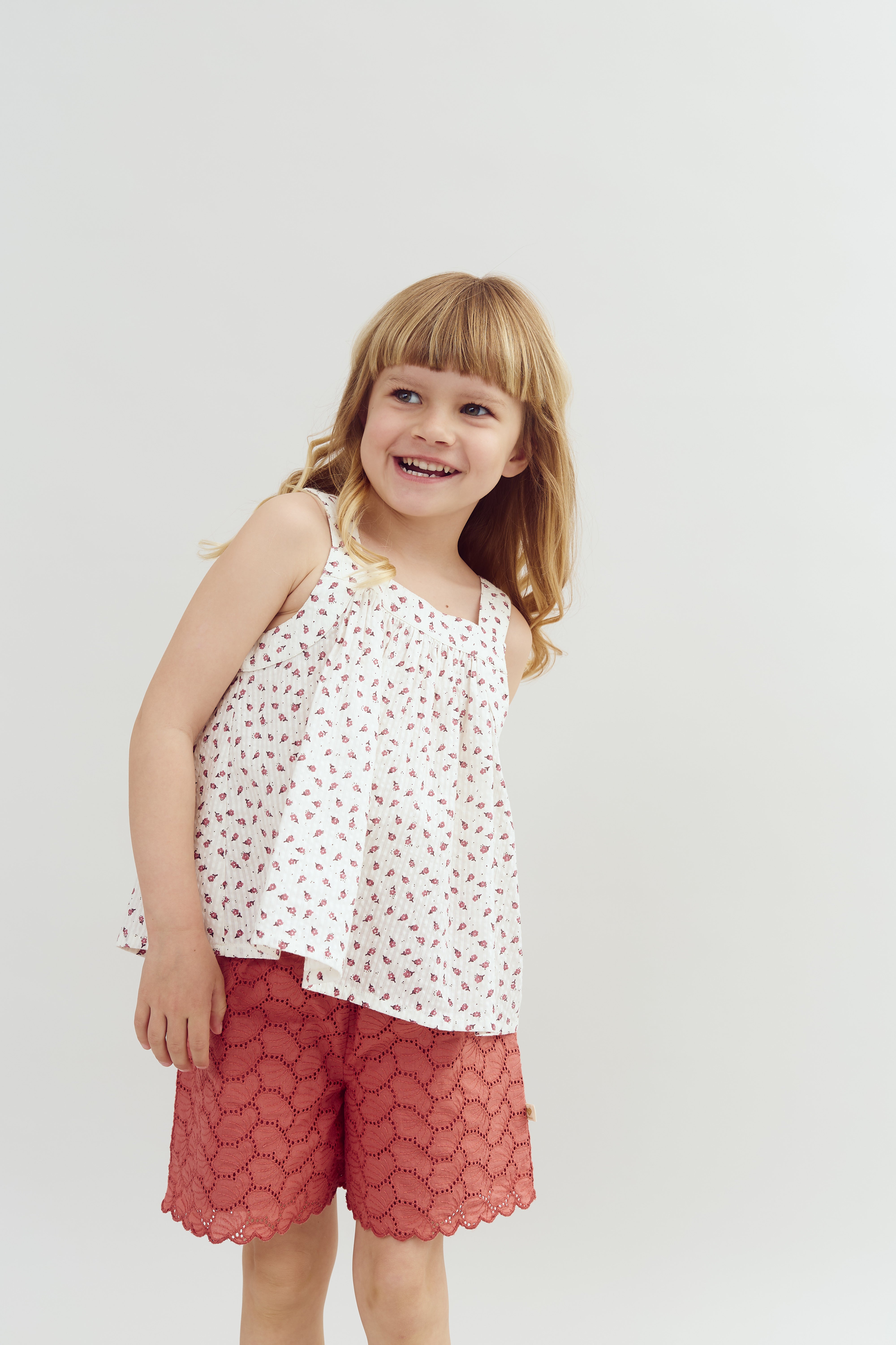 A child smiles broadly, wearing a white patterned blouse and red lace shorts. The background is plain and light-colored, enhancing the playful, joyful mood.