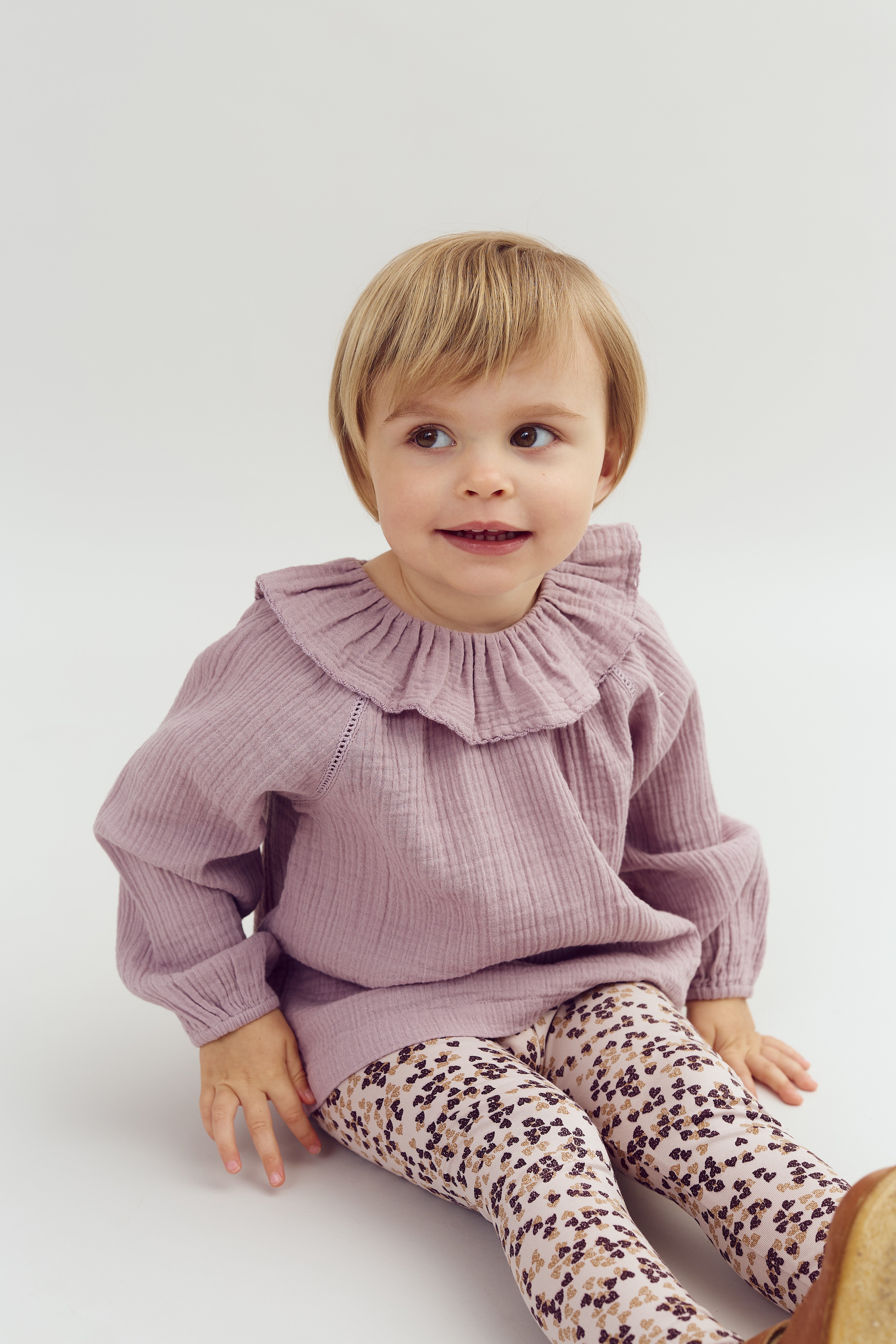 Et lille barn sidder og smiler iført en lys lilla bluse og mønstrede leggings. Baggrunden er enkle og lyse, hvilket fremhæver barnets tøj og udstråling.