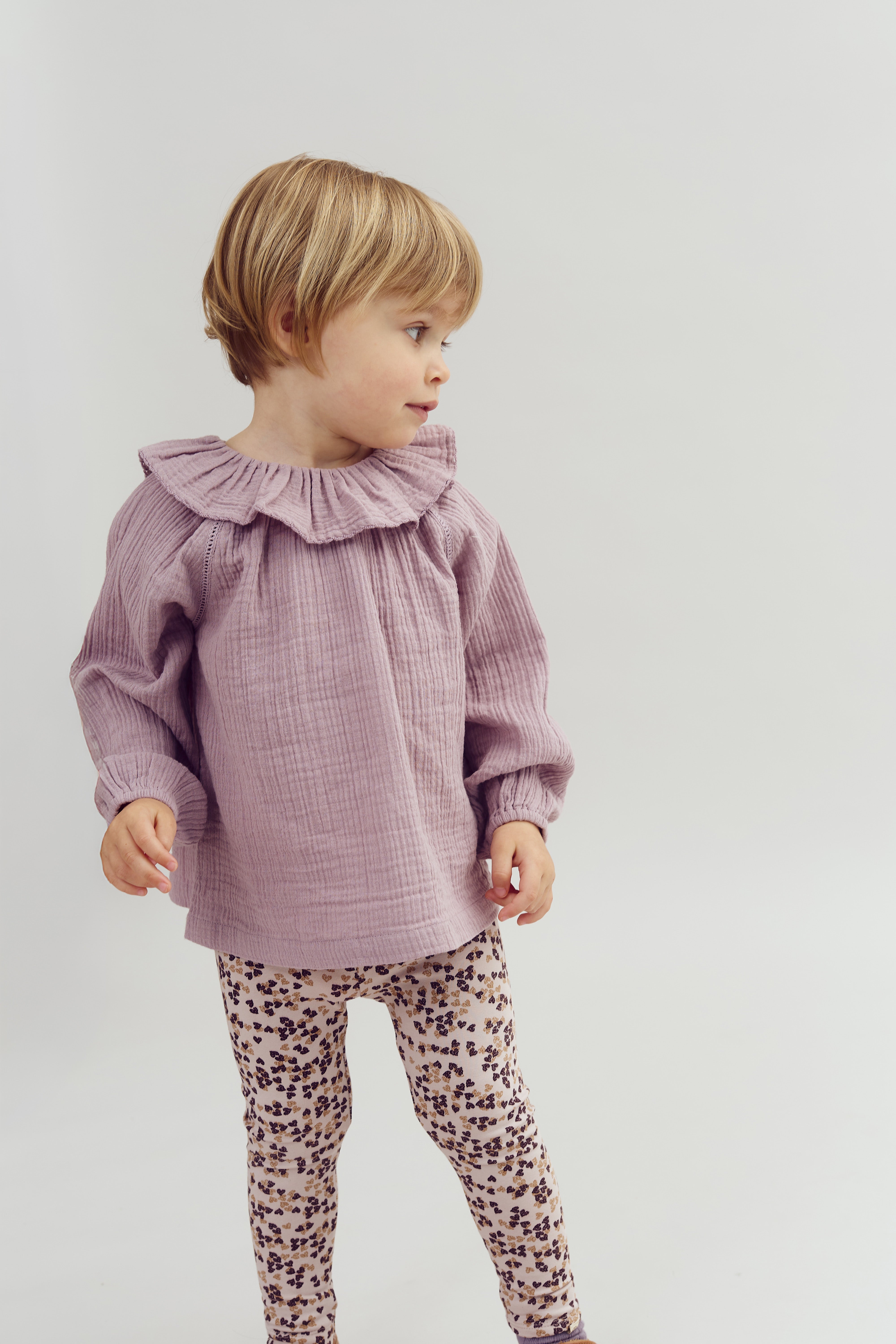 Et lille barn står iført en lilla bluse og blomstrede leggings, vender hovedet mod venstre i et neutralt, lyst rum.