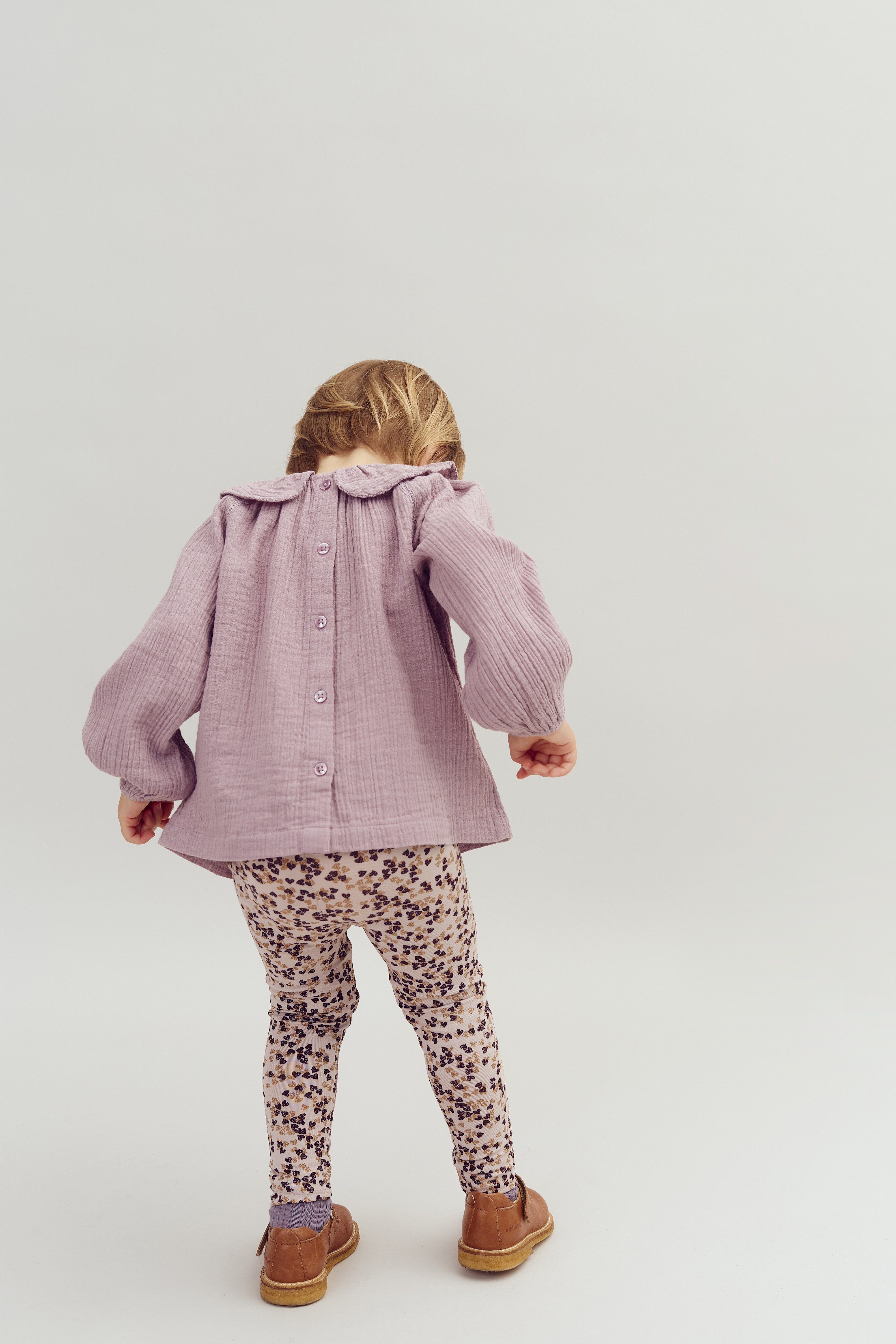 Et barn iført en lilla bluse og mønstrede leggings står med ryggen til, bøjet let forover, i et lyst, neutralt rum.