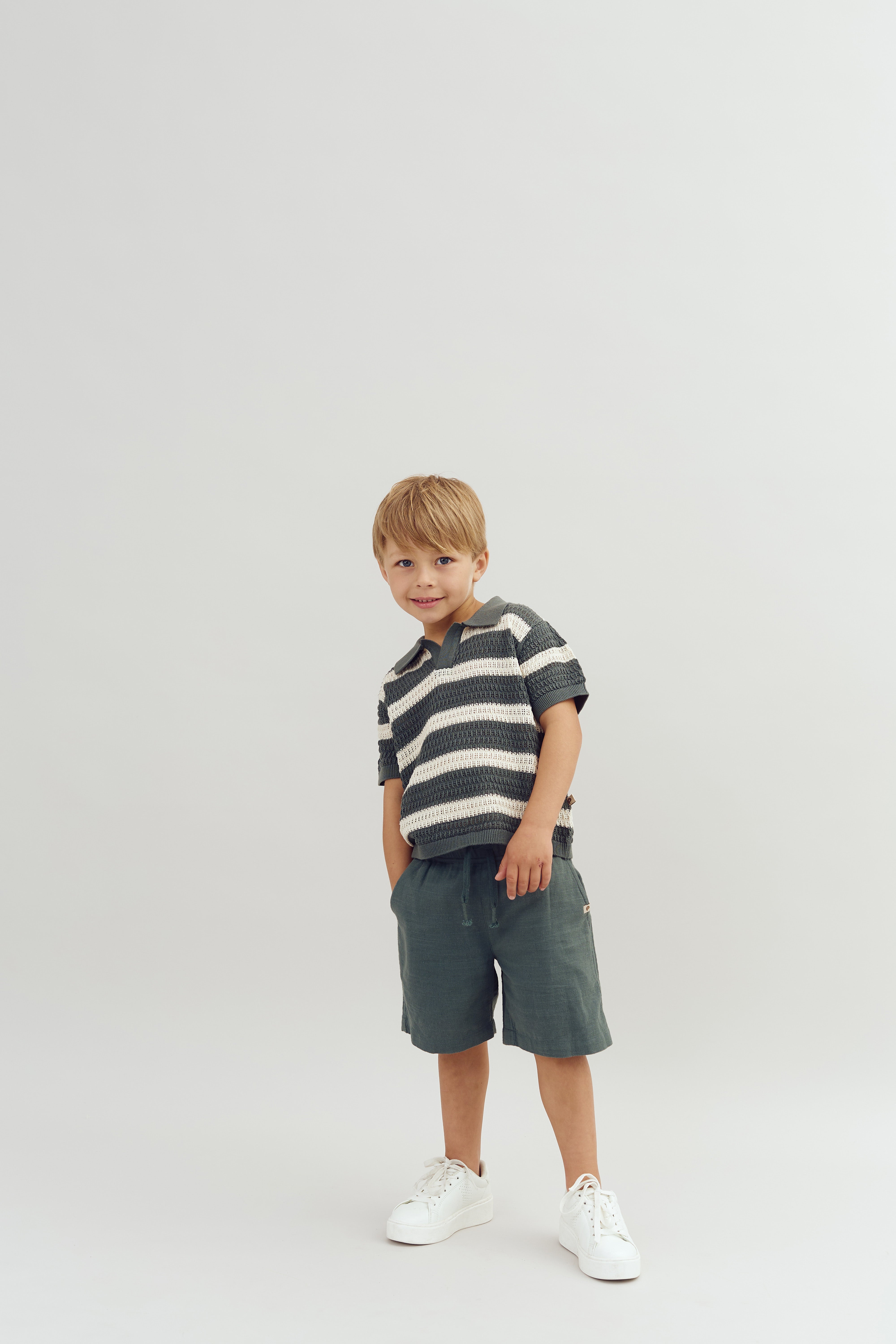 A young child stands smiling, wearing a striped shirt, green shorts, and white sneakers against a plain white background.