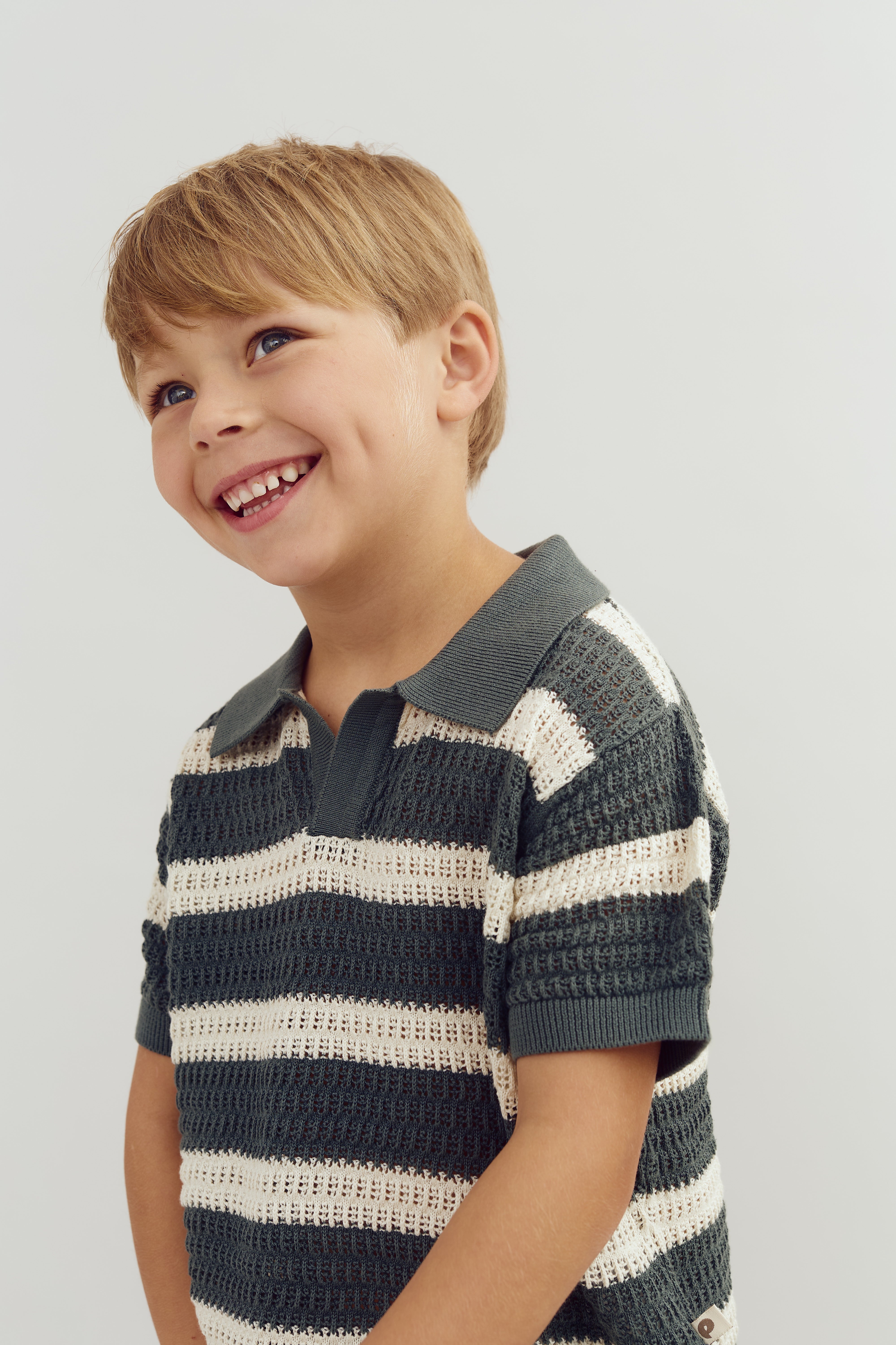 A young boy smiles, showing dimples, dressed in a striped collared sweater, standing against a plain light background.