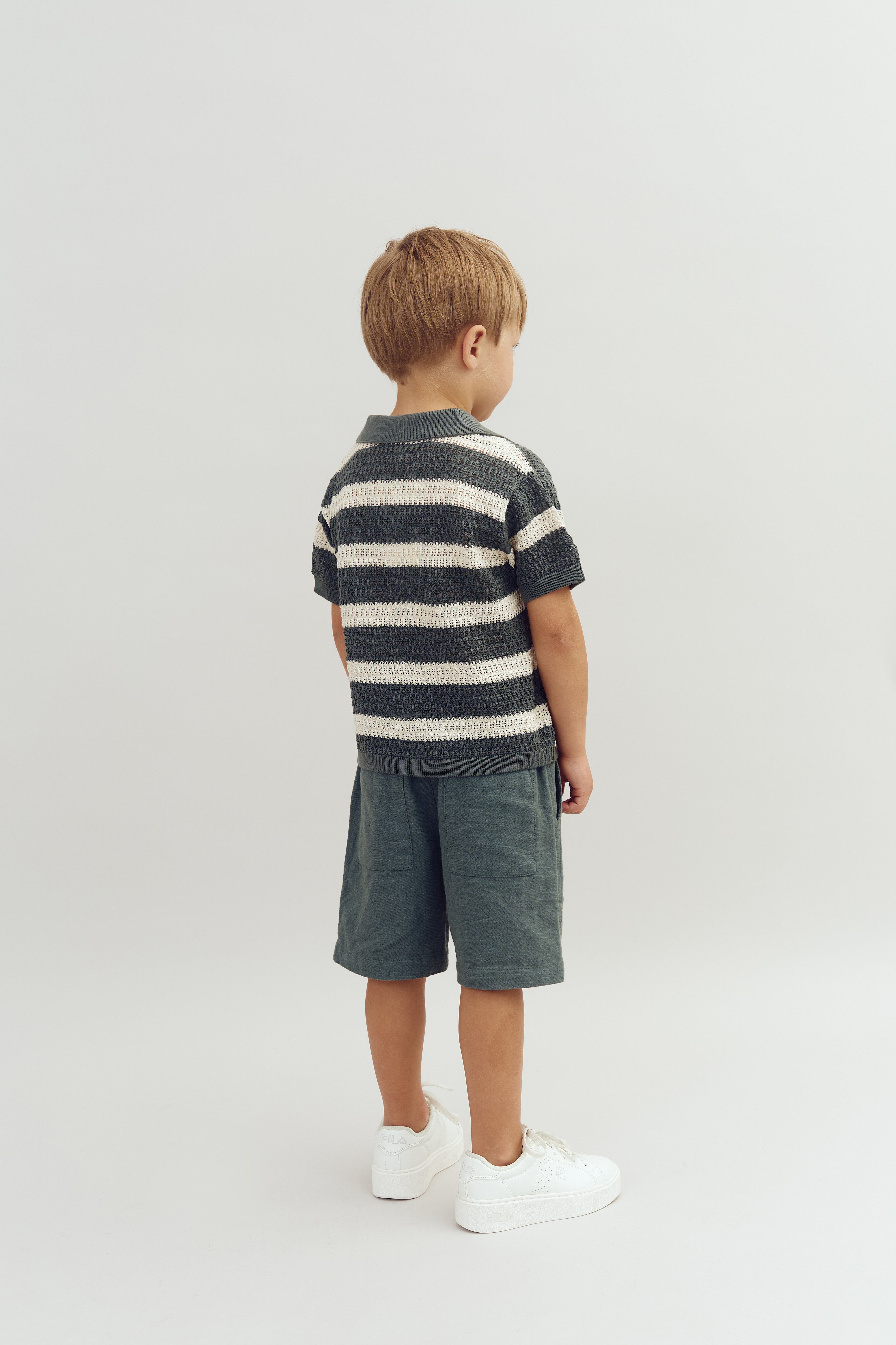 A child stands with back facing, wearing a striped shirt, green shorts, and white sneakers in a plain, well-lit setting.