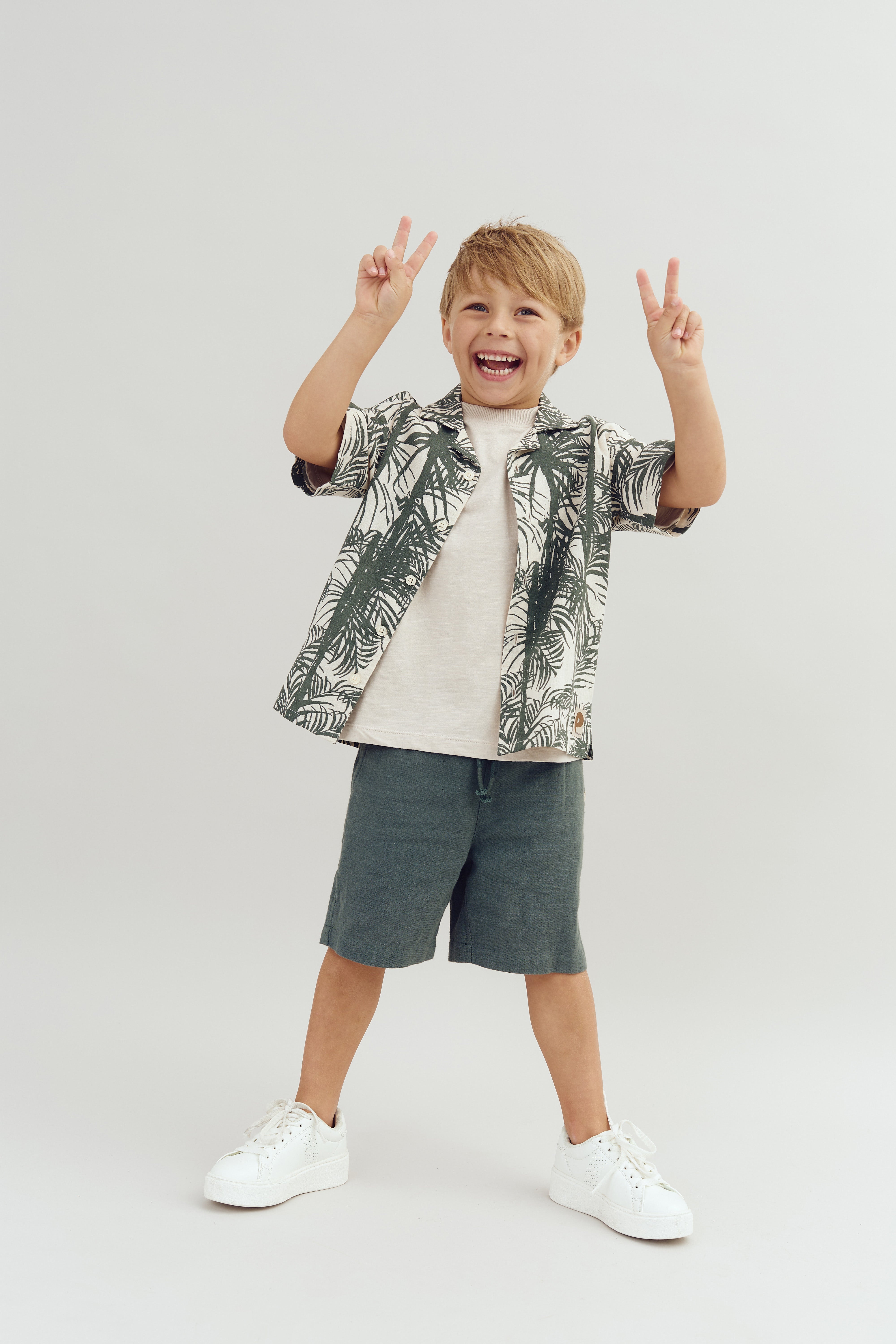 Dreng smiler og holder to sejrstegn med hænderne. Han har en skjorte med palmemønster, shorts og sneakers på. Baggrunden er en neutral, lys farve.