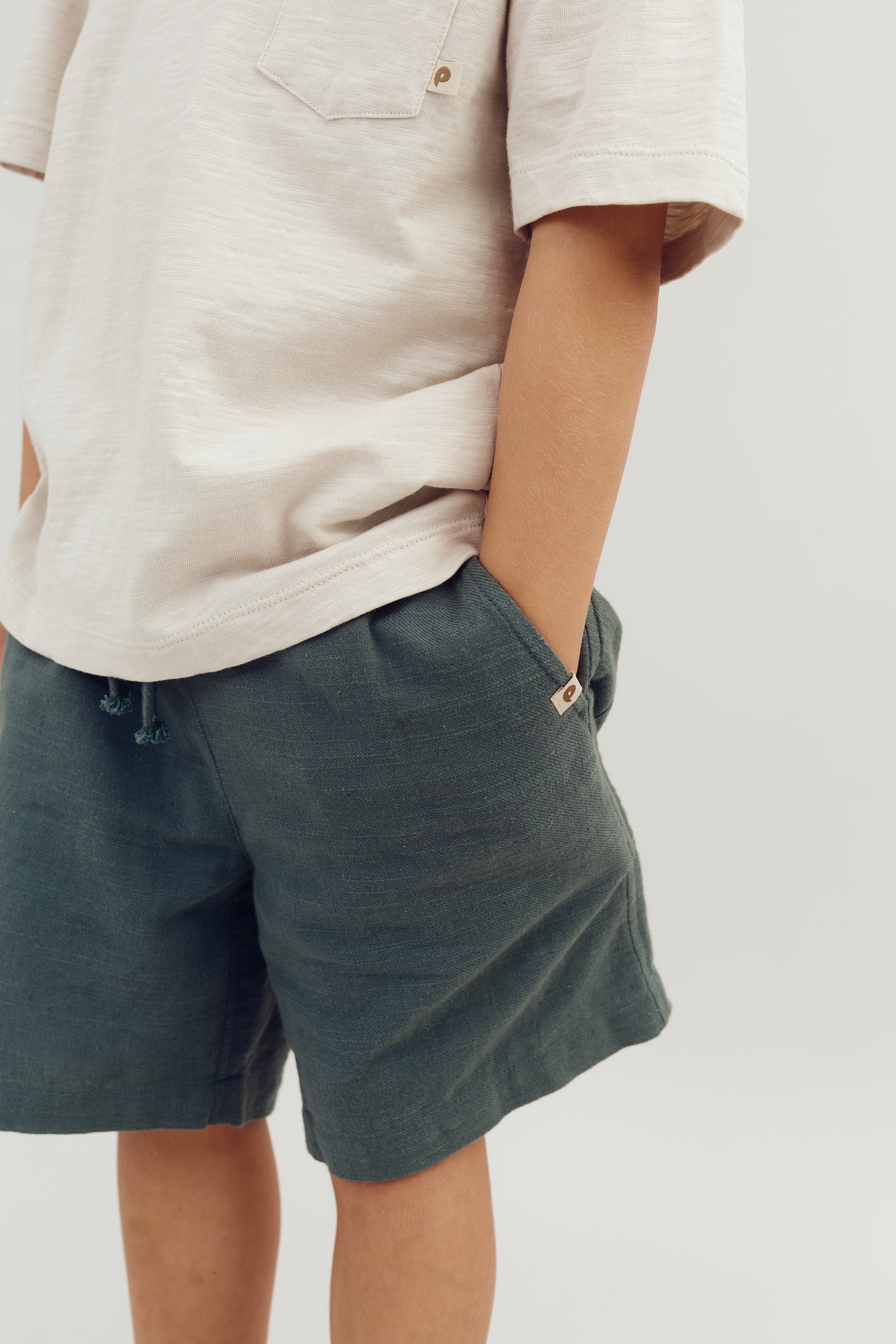 En person iført en lysegrå t-shirt med lomme og mørkegrønne shorts har hænderne i lommerne i en neutral indendørs baggrund.