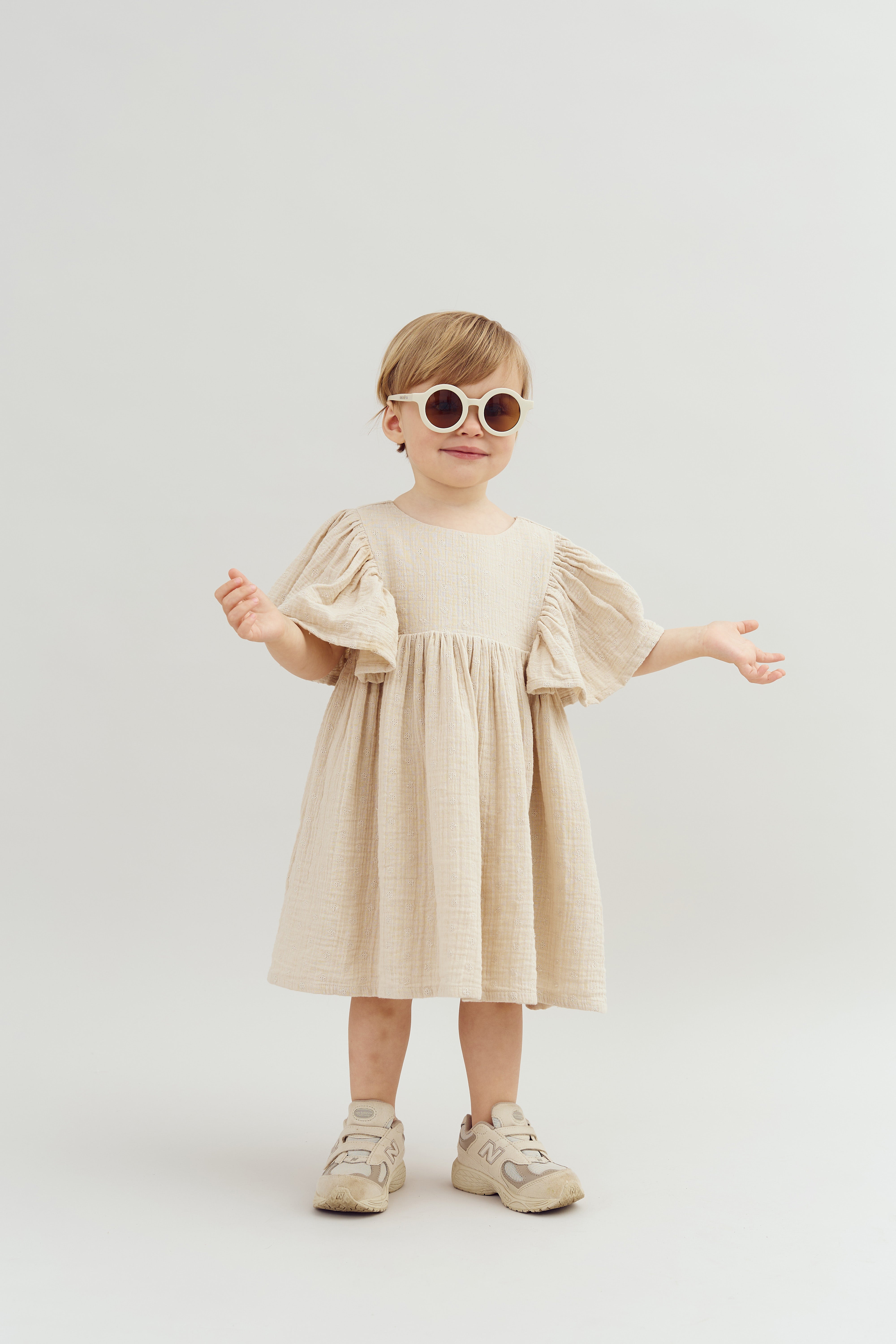 A child stands with arms outstretched, wearing round sunglasses, a cream-colored dress with puffy sleeves, and beige sneakers, set against a plain light background.