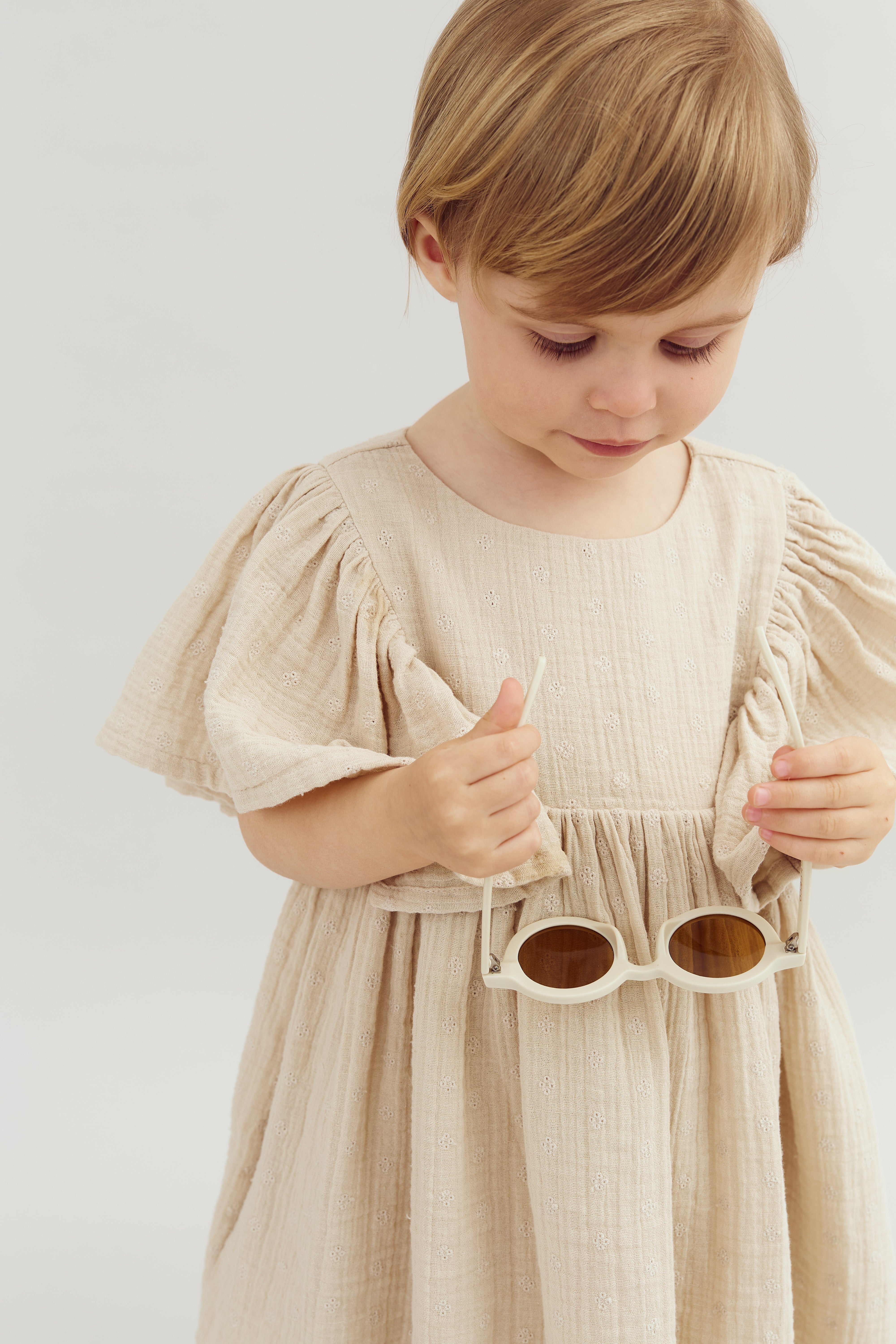 A child, wearing a beige, textured dress with puffed sleeves, holds a pair of beige sunglasses. The background is plain and light-colored, emphasizing the child’s attire and focus.