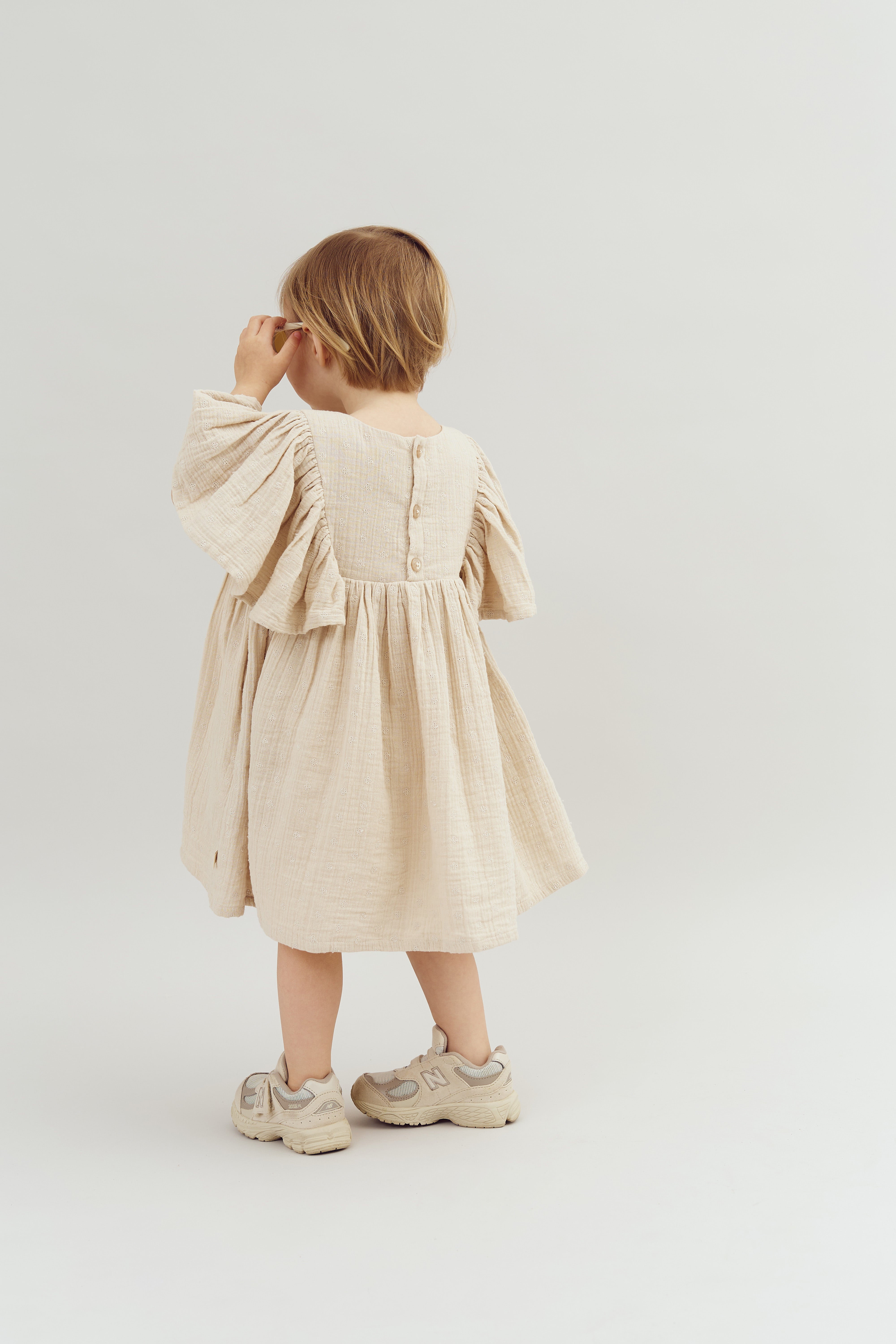 A child wearing a cream-colored dress with buttons and beige sneakers stands facing away, holding something near their face, against a plain white background.