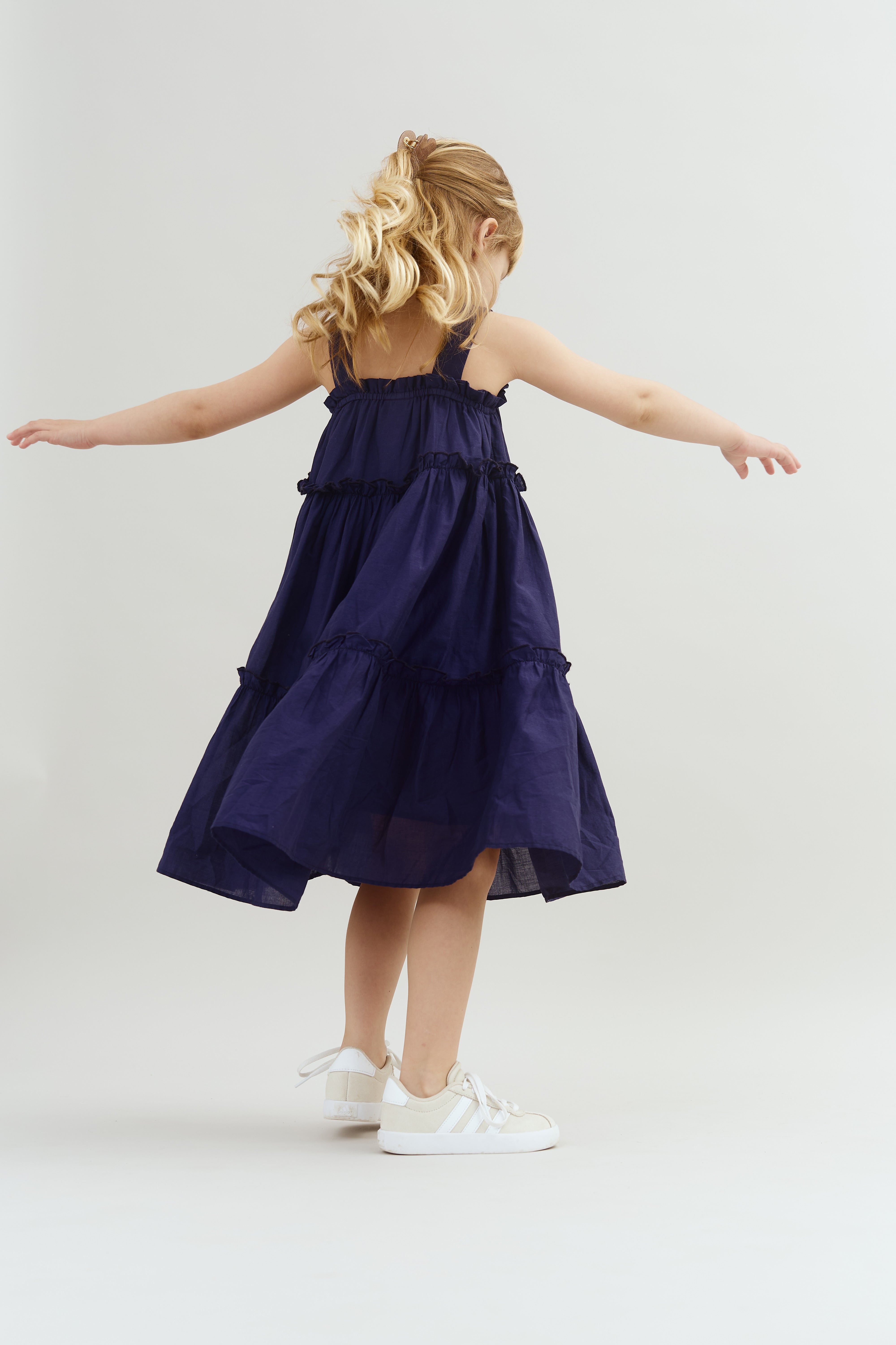 A child wearing a navy dress and white sneakers twirls playfully, arms outstretched, against a plain light grey background.